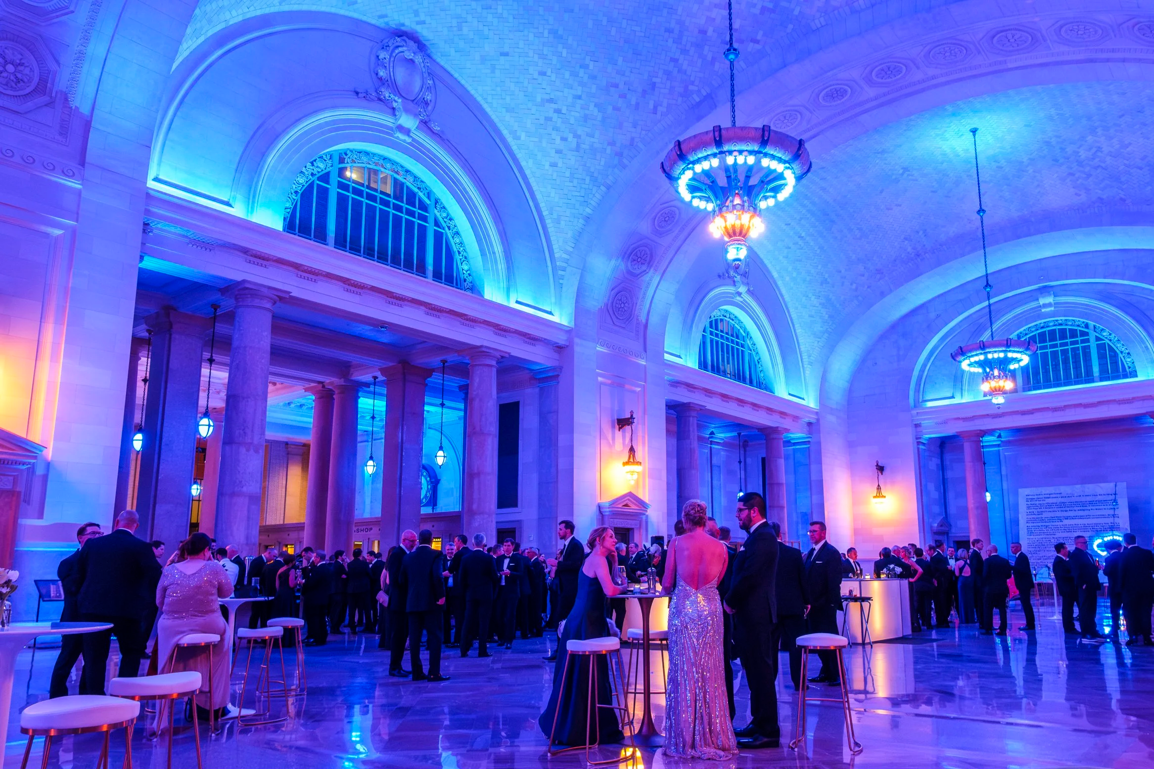 The interior of Michigan Central Station with blue, purple and gold lighting illuminates a room of people wearing suits and gowns.