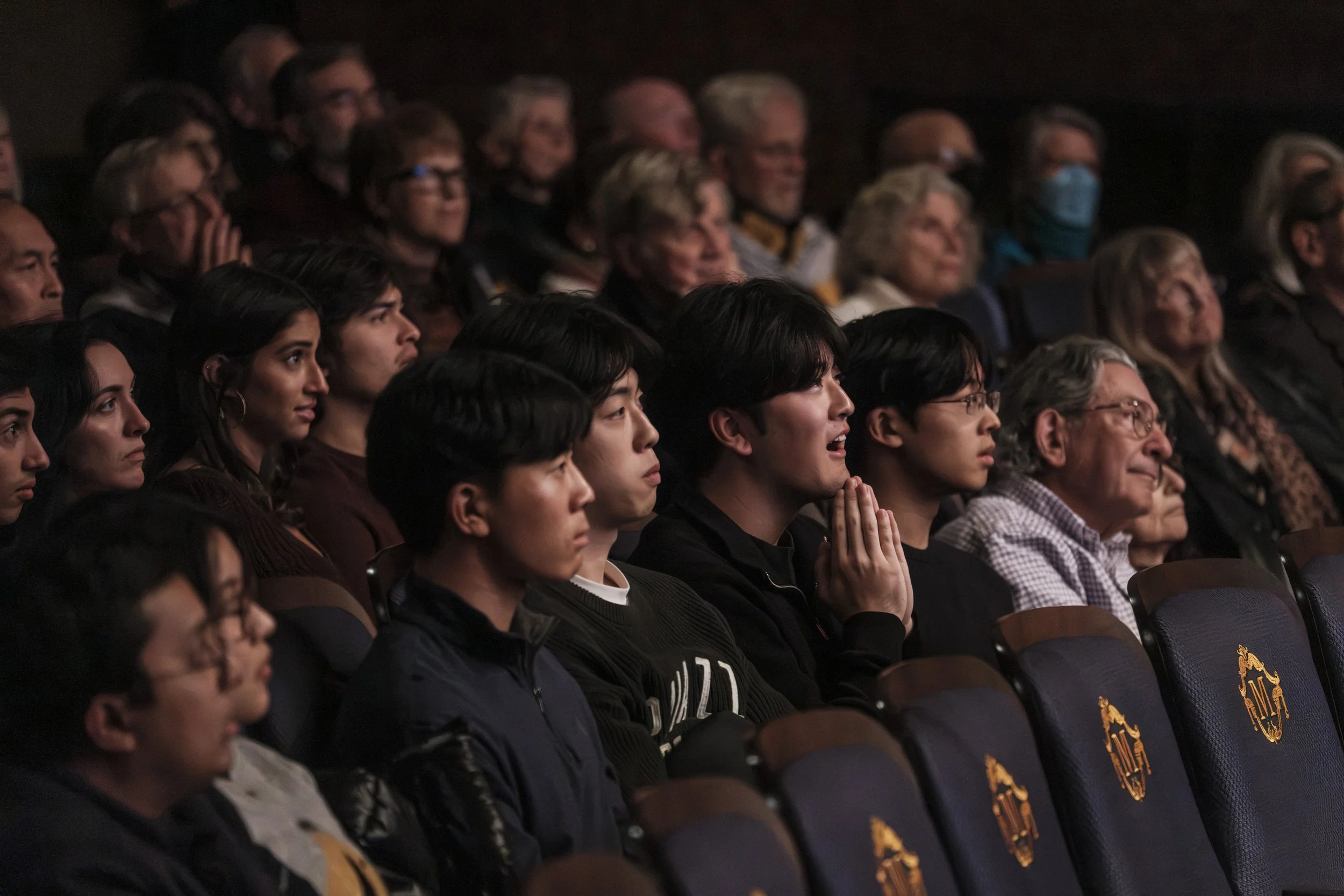 Young men in an audience listen to an orchestra performance while one of the men in the group holds his hands to his mouth in astonishment.