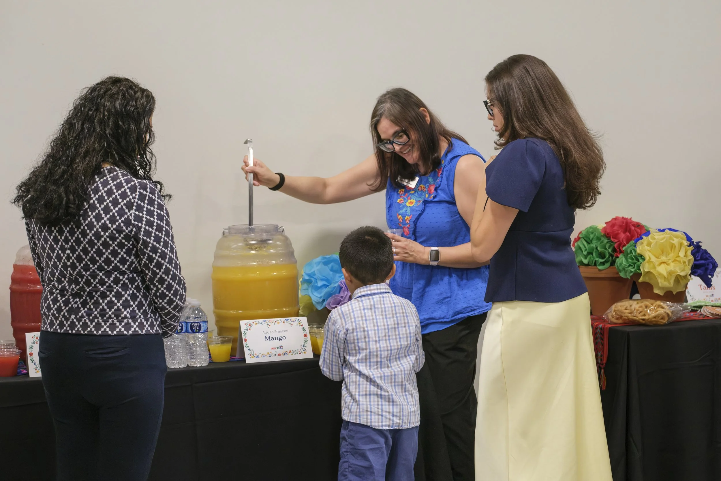 A woman smiles and holds a ladle while serving mango juice to a family with a young boy.