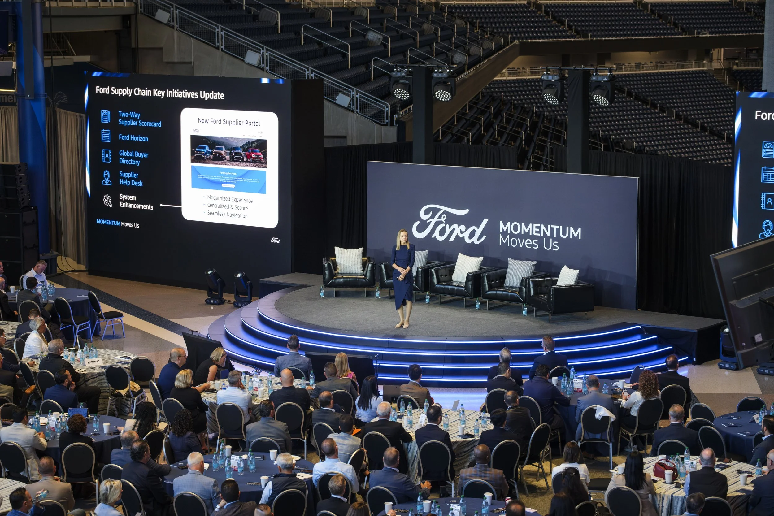 A woman stands in a dress on a large stage with a banner behind that reads "Ford Momentum Moves Us" while a crowd of people sit at tables that are placed around the stage.
