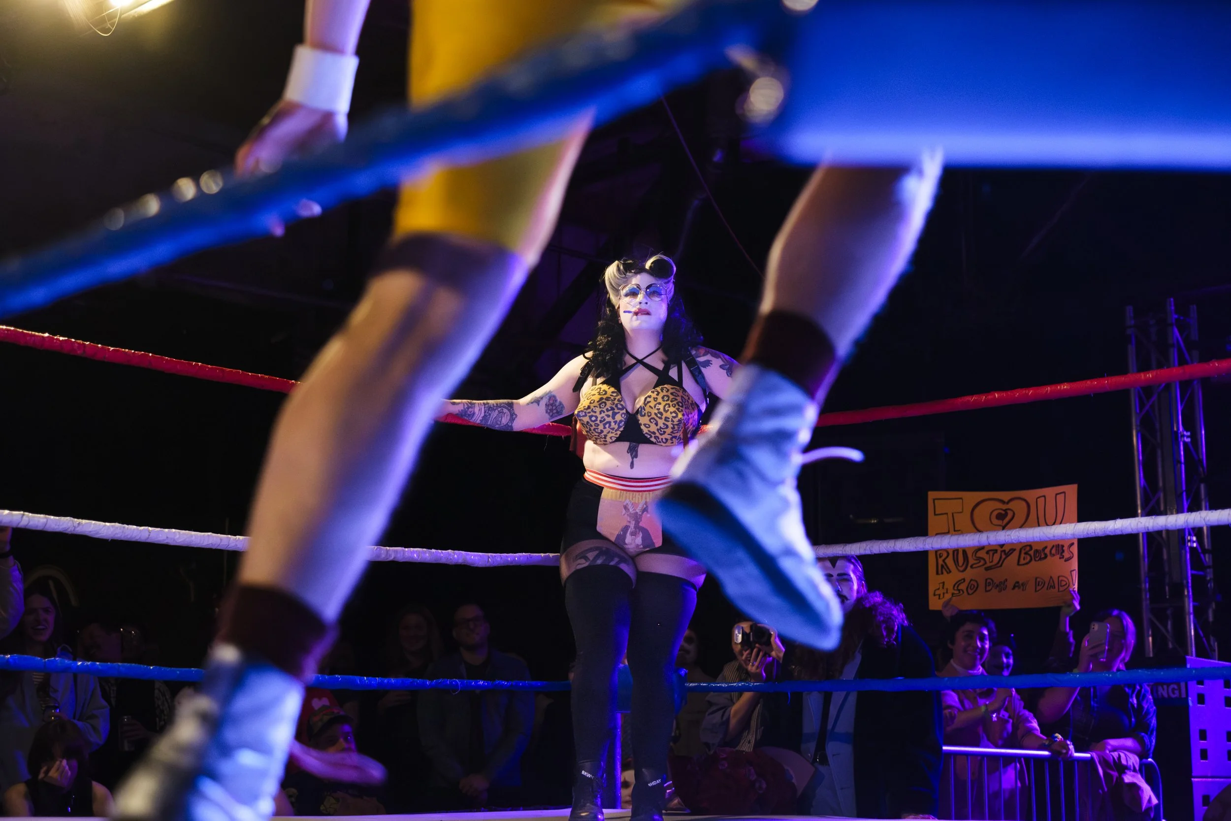 A clown wrestler, known as Rusty Busches, stares at her opponent, framed between the legs of her opponent.