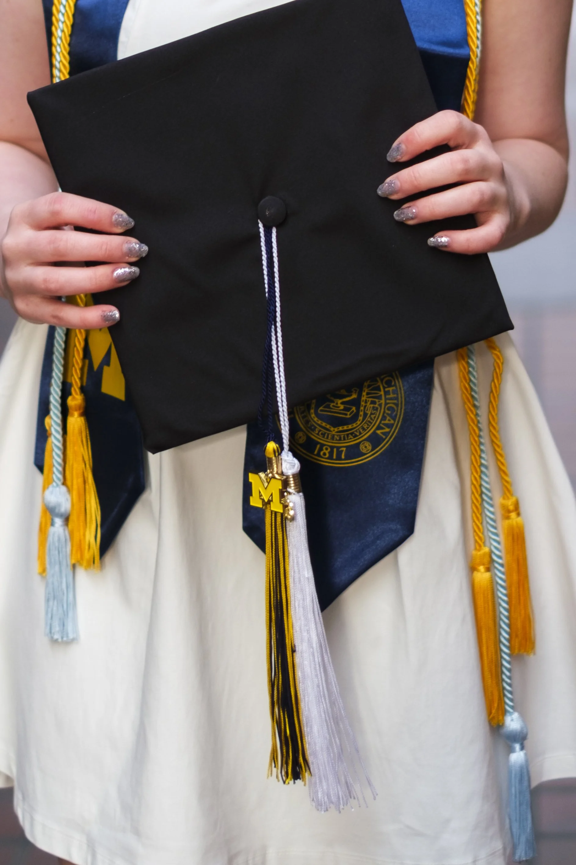 Hands with sparkly nail polish hold a graduation cap with tassels featuring a Block M.