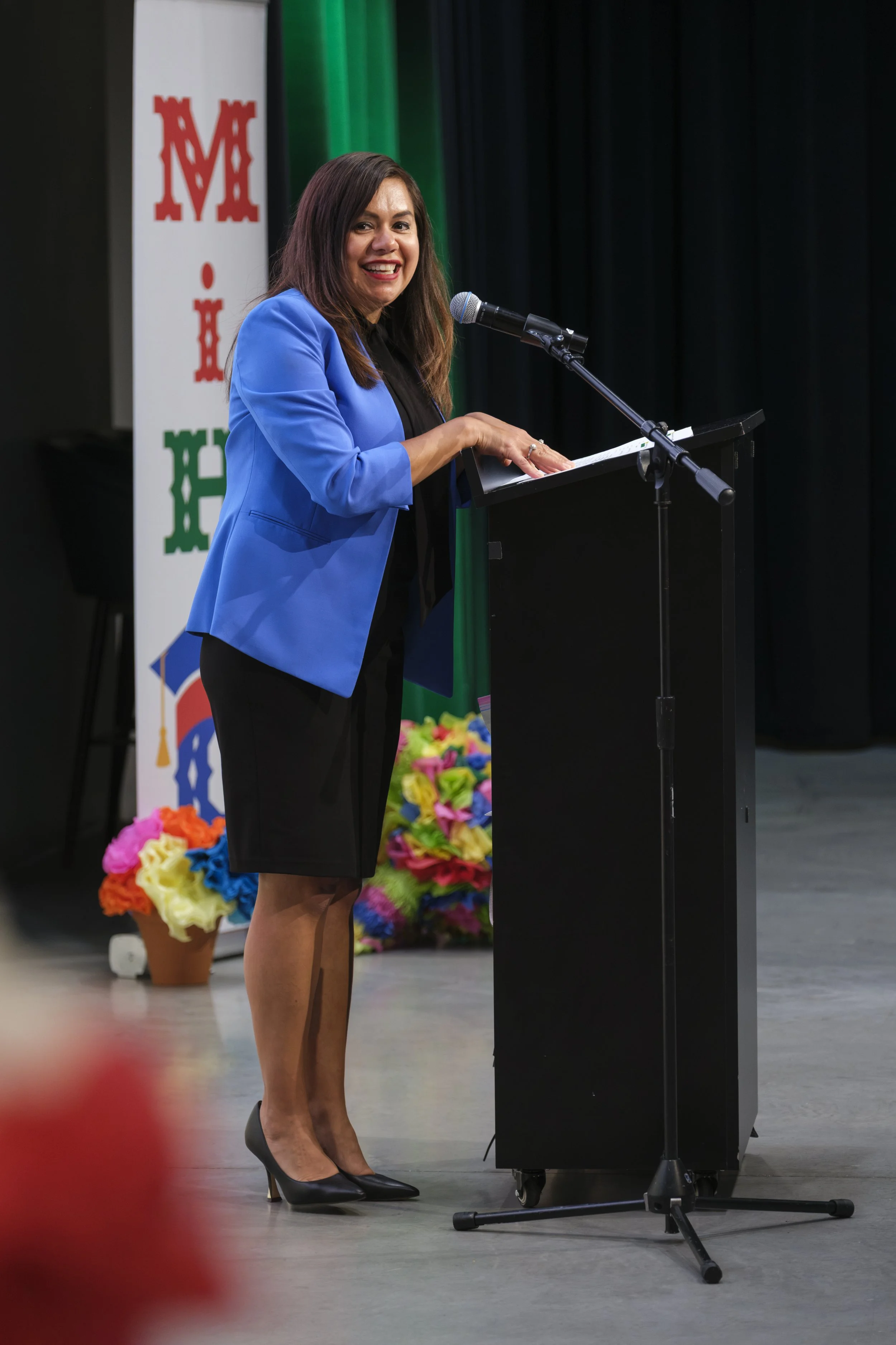 A young woman in a blue blazer stands and smiles while speaking into a microphone behind a podium.