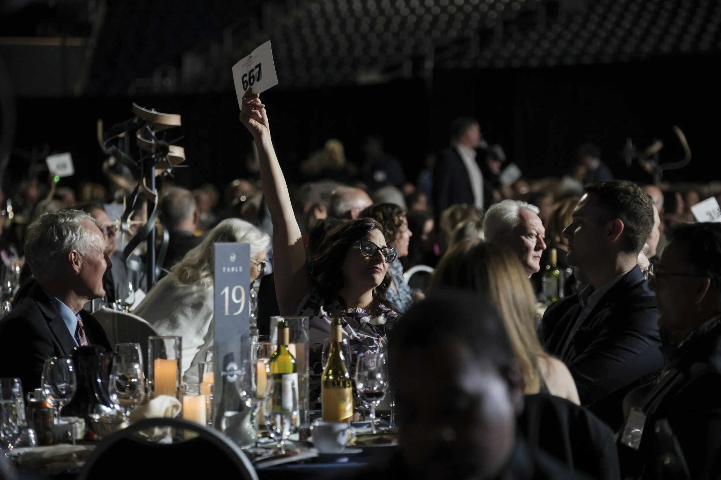 A woman partially illuminated by stage lighting holds up a paper with a number while sitting at a table.