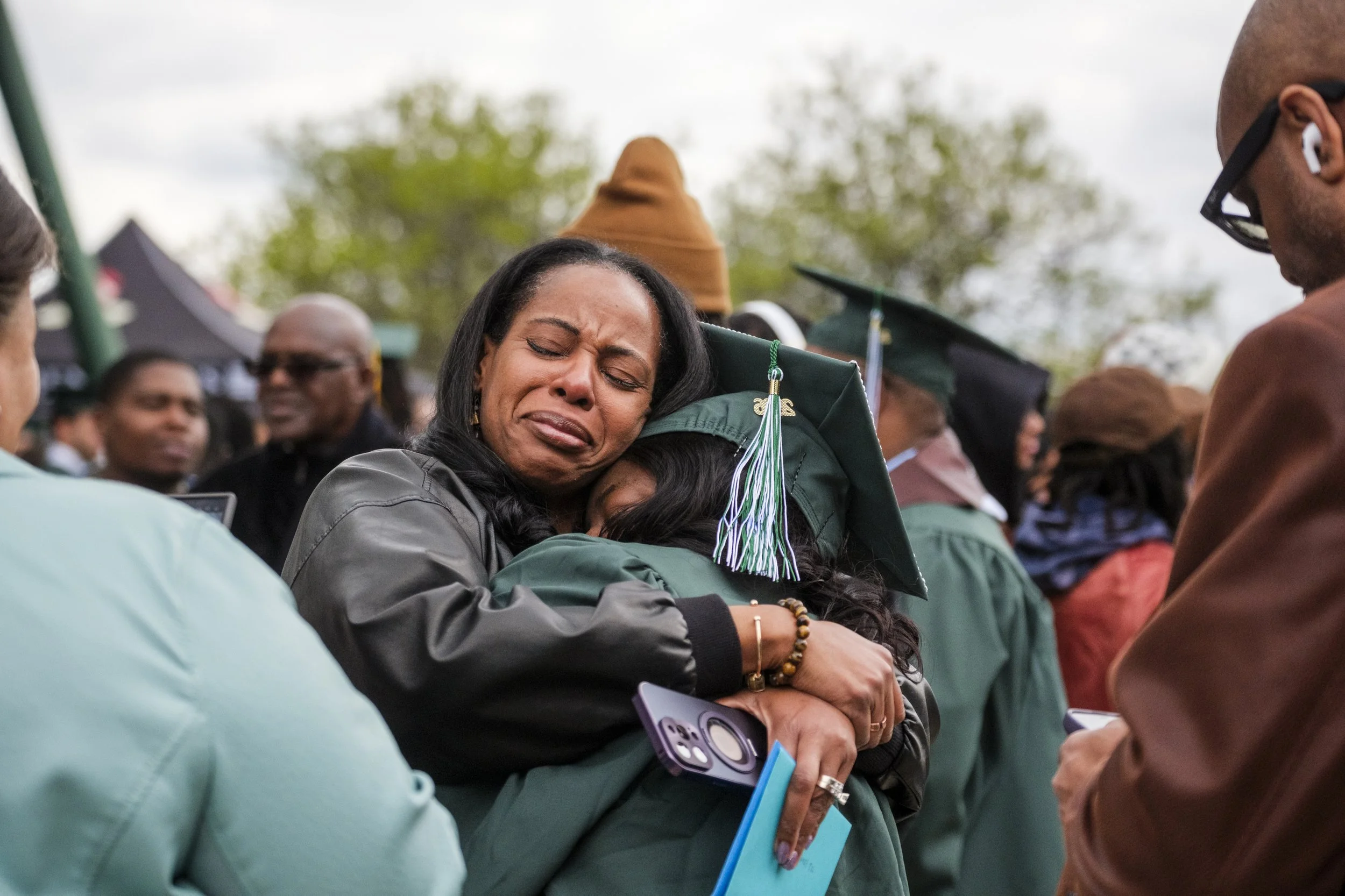 A person smiles while crying tears of joy while hugging their daughter, wearing a green graduation cap, gown and tassel.
