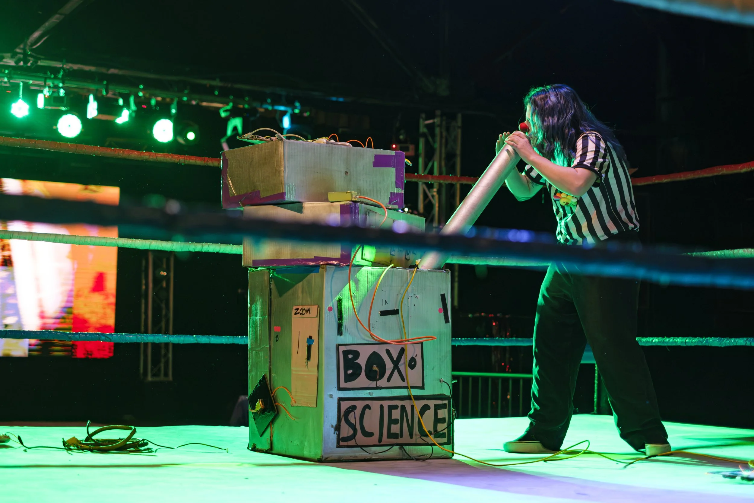 A referee in clown make-up looks into a prop with the words "Box Science" on the outside of it, while standing in a wrestling ring.