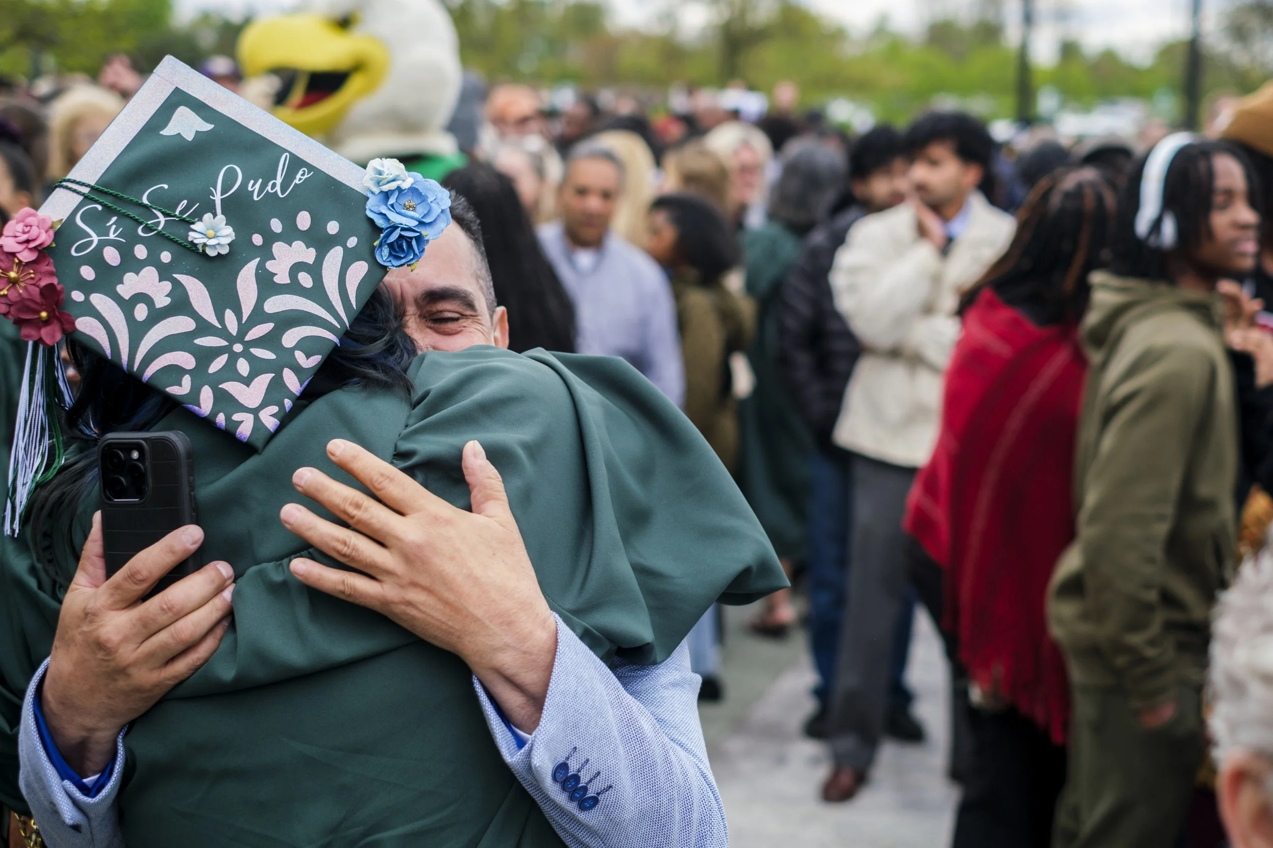 A man embraces a graduate with a decorated graduation cap that reads "Si Se Pudo" with a crowd of people in the background.