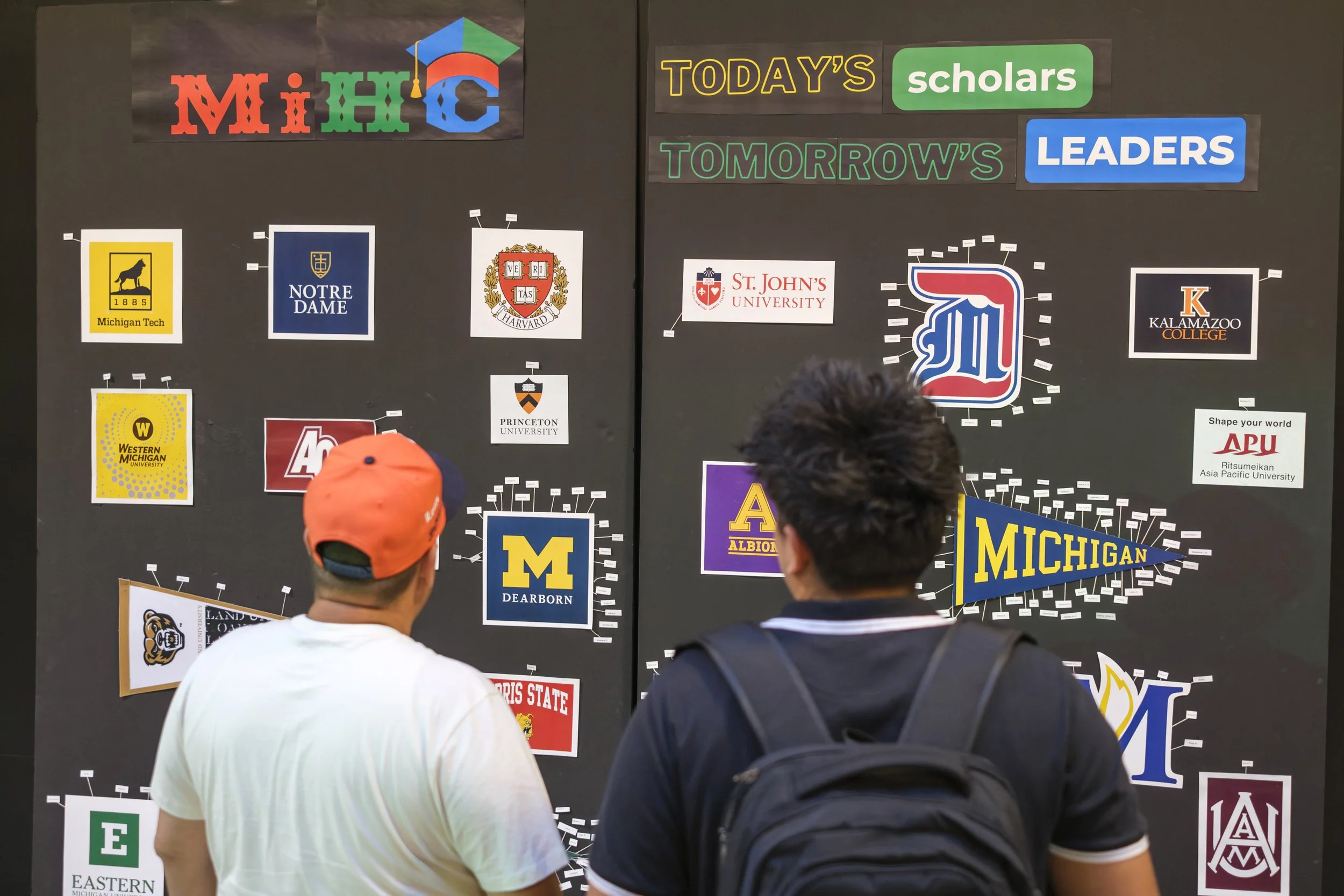 A board with college and university logos and names attached to them stands while two people stand in front of it, looking at the board.