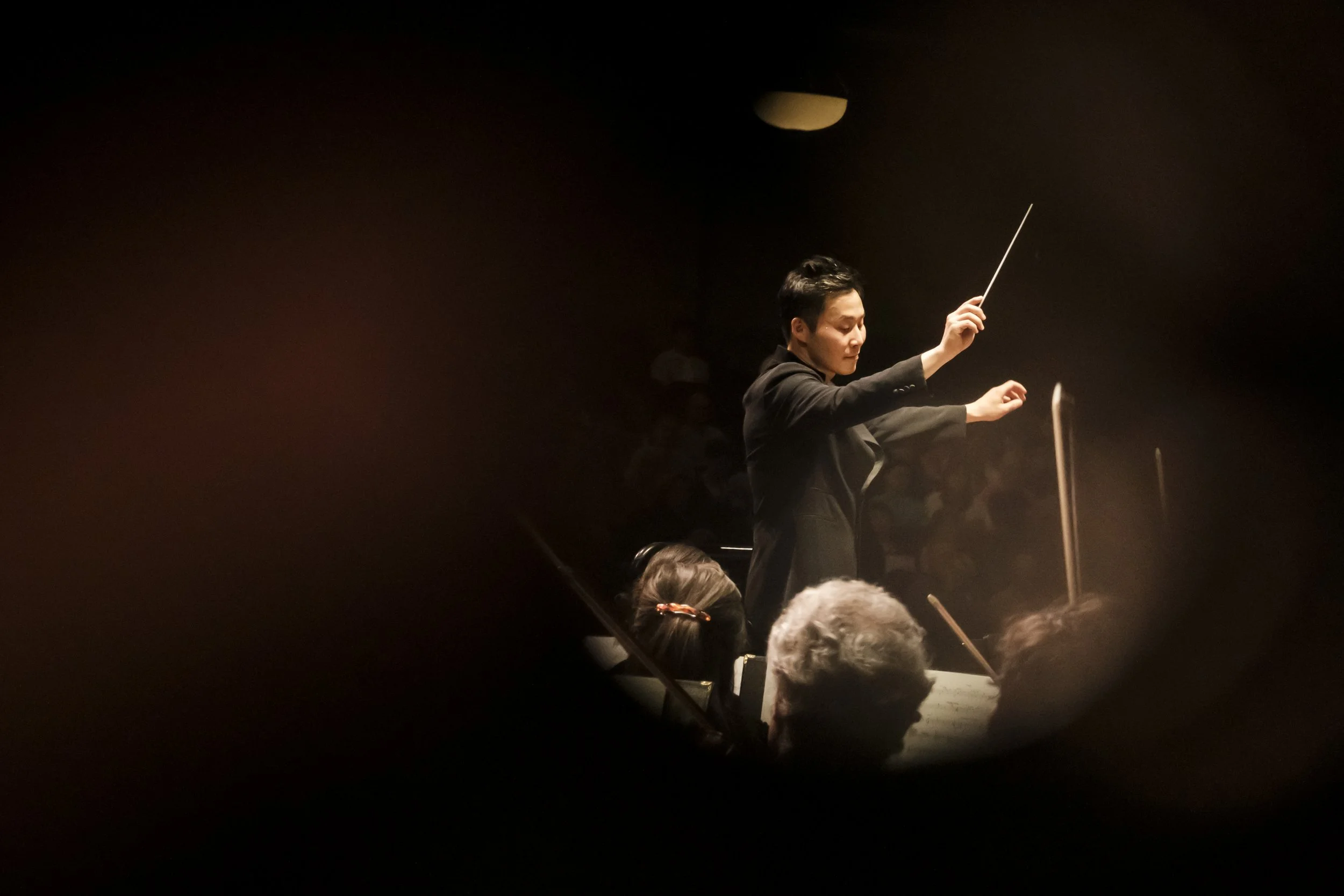 Conductor Earl Lee stands on stage directing the orchestra, framed in a round window.