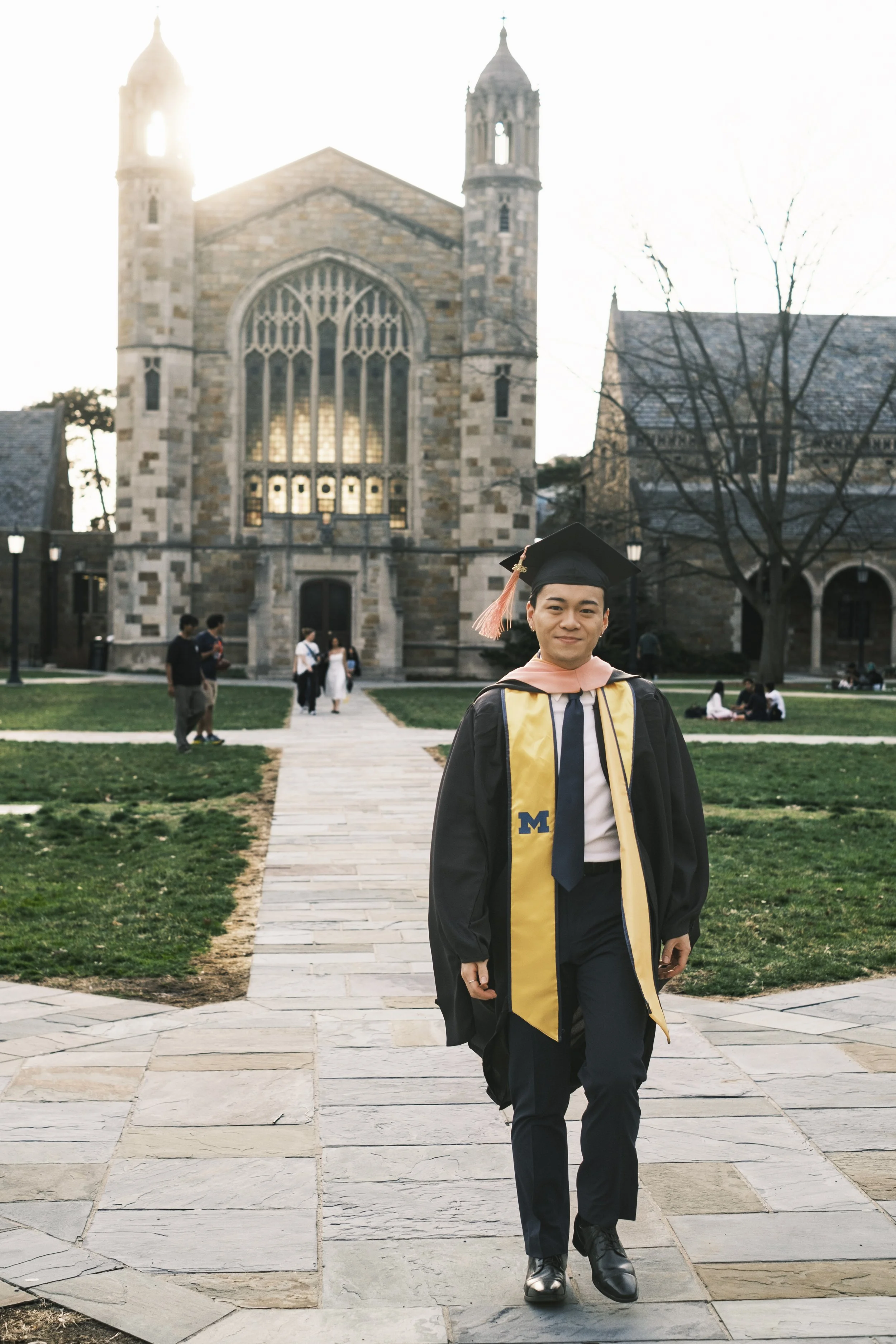 A person smiles while walking down a brick walkway wearing a graduation cap and gown with a maize colored sash with a historic building in the background.