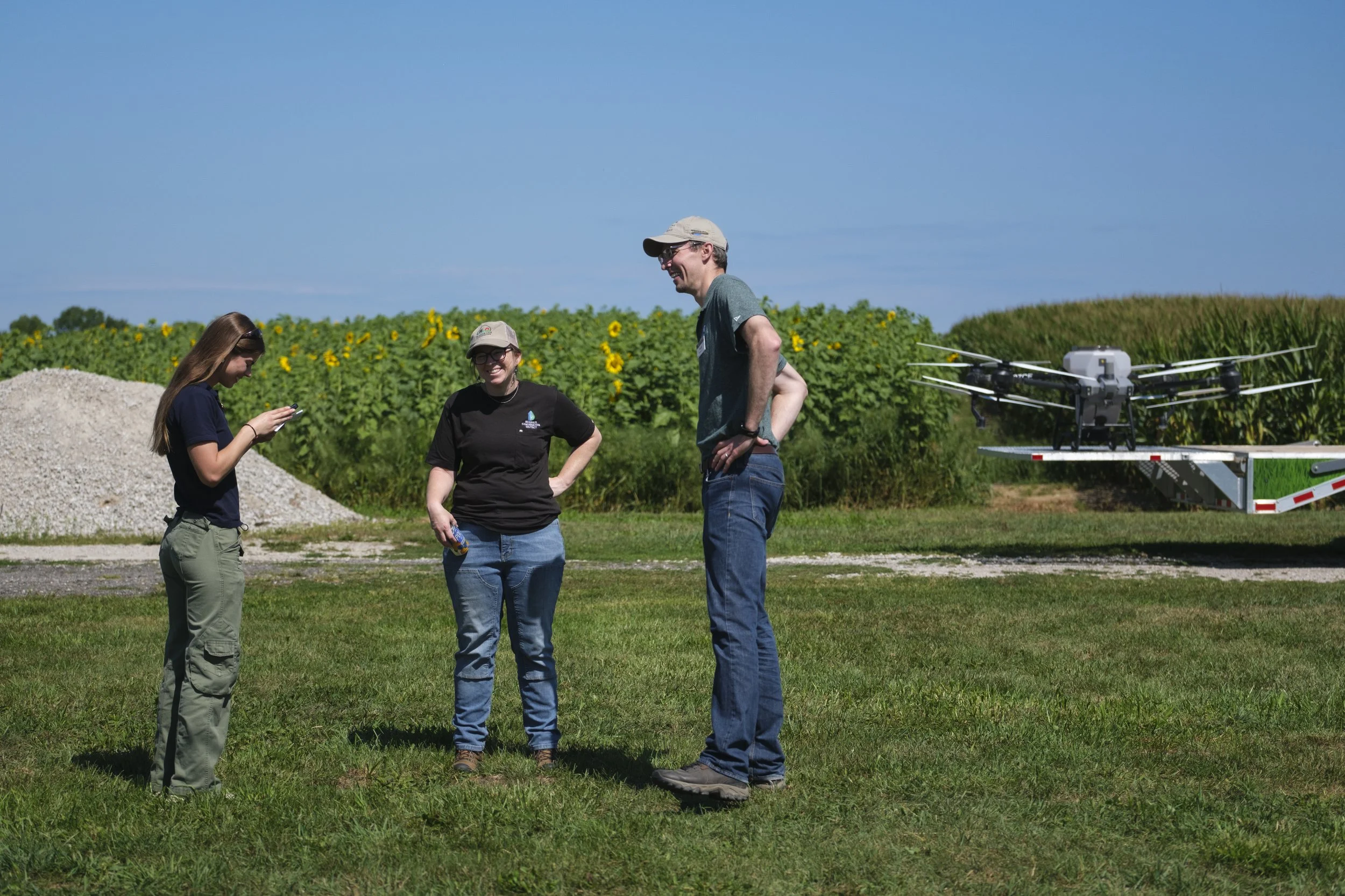 A group of three people stand in a field smiling at one another in front of a field of sunflowers with a drone sitting on a flat bed truck in the background.