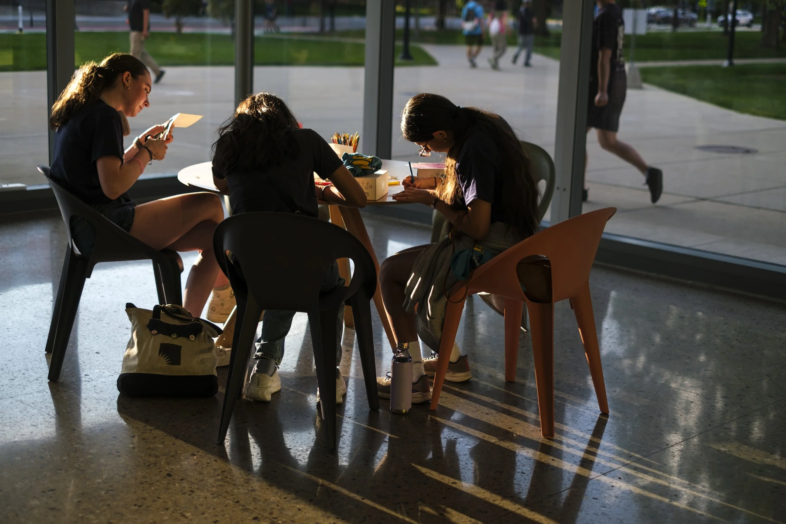 A group of three students sit in a room using scissors and coloring while sitting at a table with golden sunlight coming through the window.