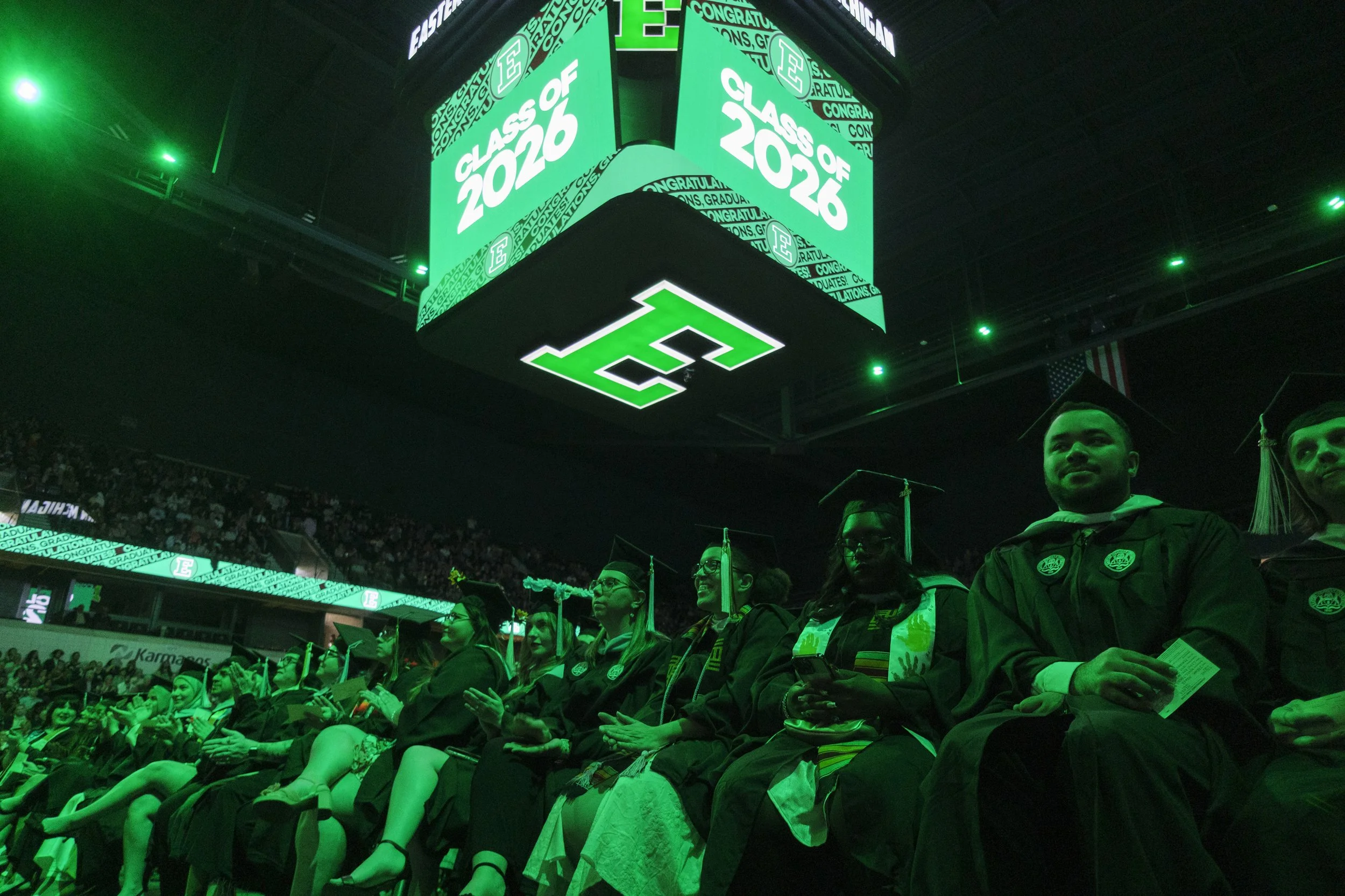 People sit in an auditorium lit with green lights with a jumbo tron that reads "class of 2026" and a block E on the bottom.