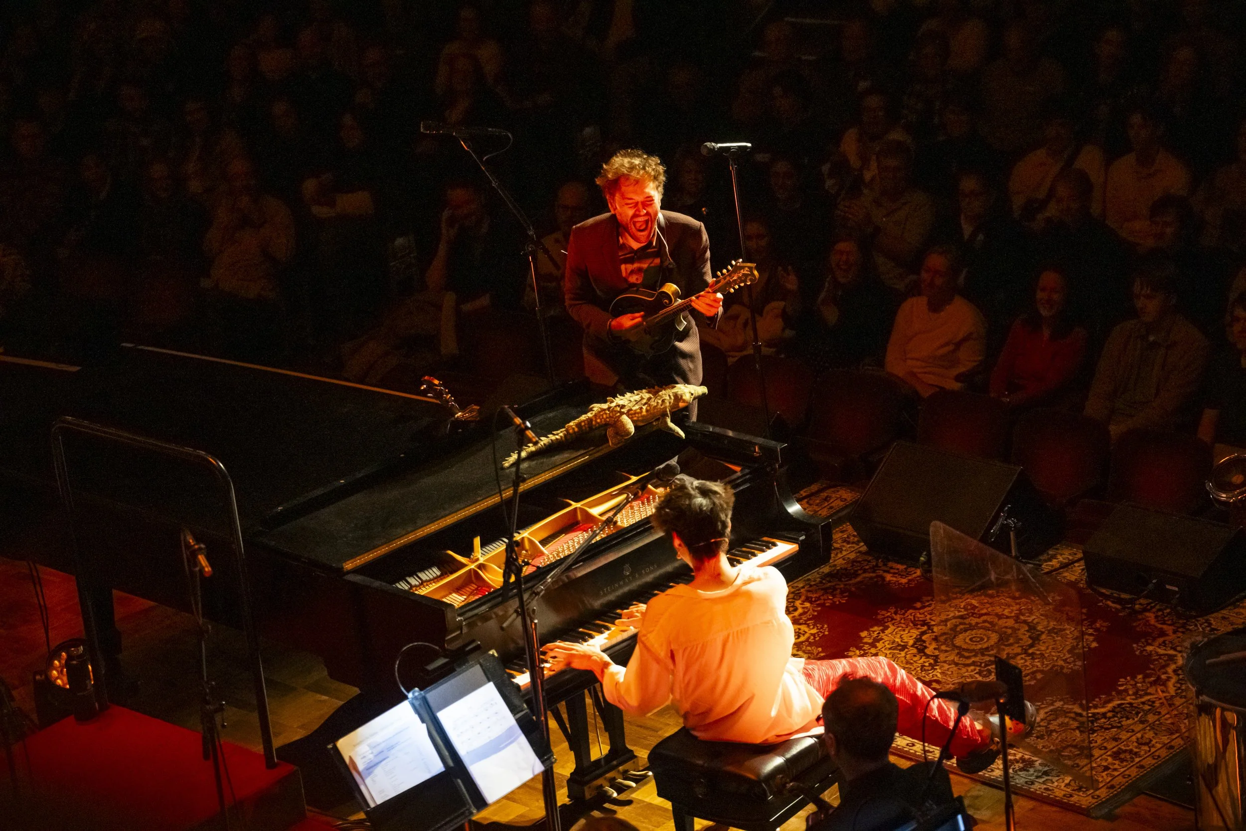 Musician Chris Thile yells on stage, illuminated by red and orange lighting, while facing musician Jacob Collier, during a concert.