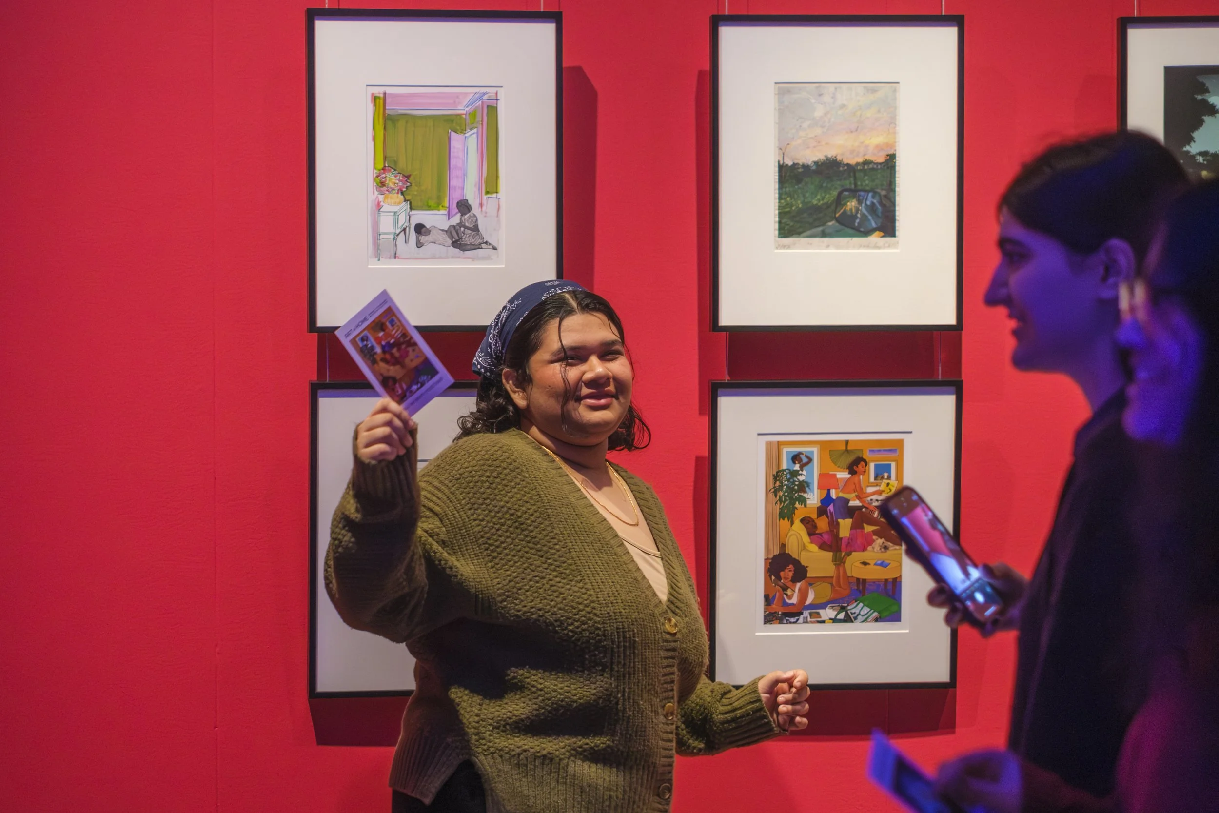A student holds up a postcard while smiling in front of a wall of framed artwork.