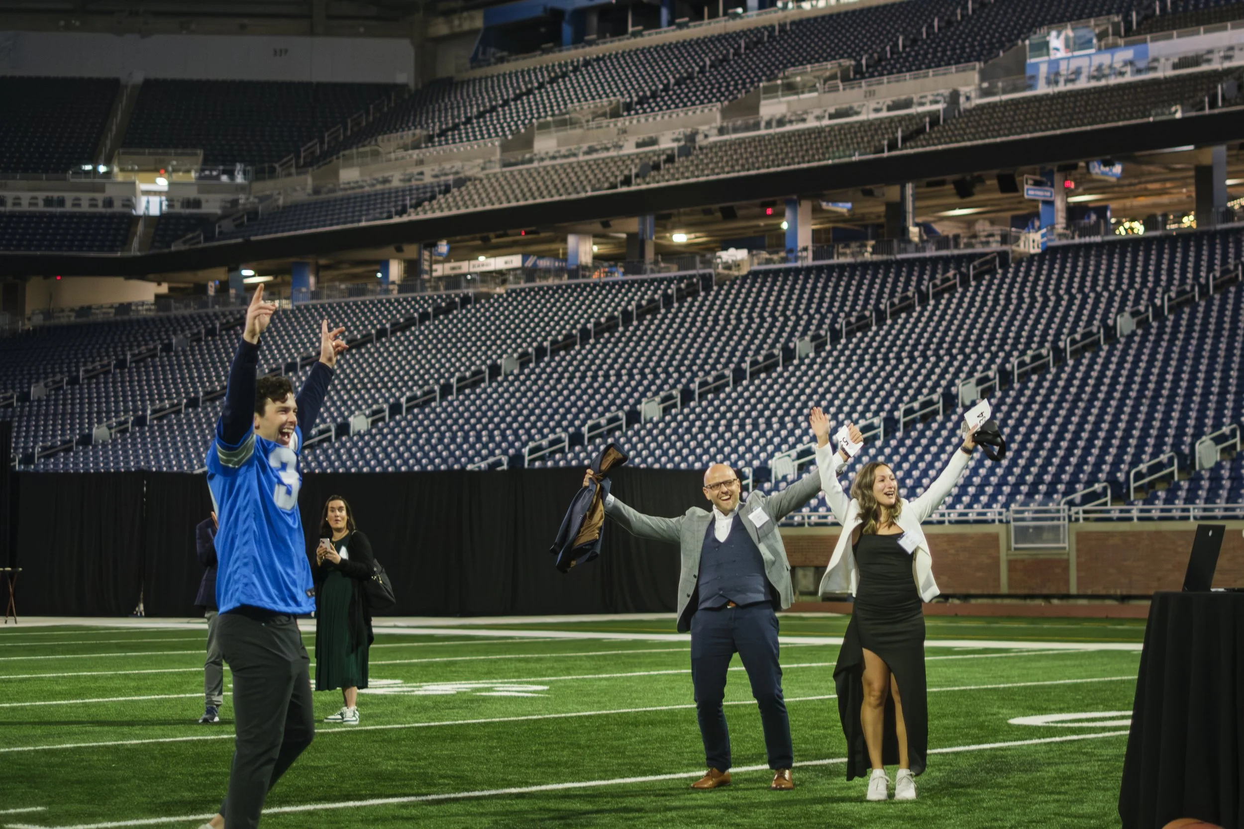 Three people standing on football turf throw their arms in the air with stadium seats in the background.