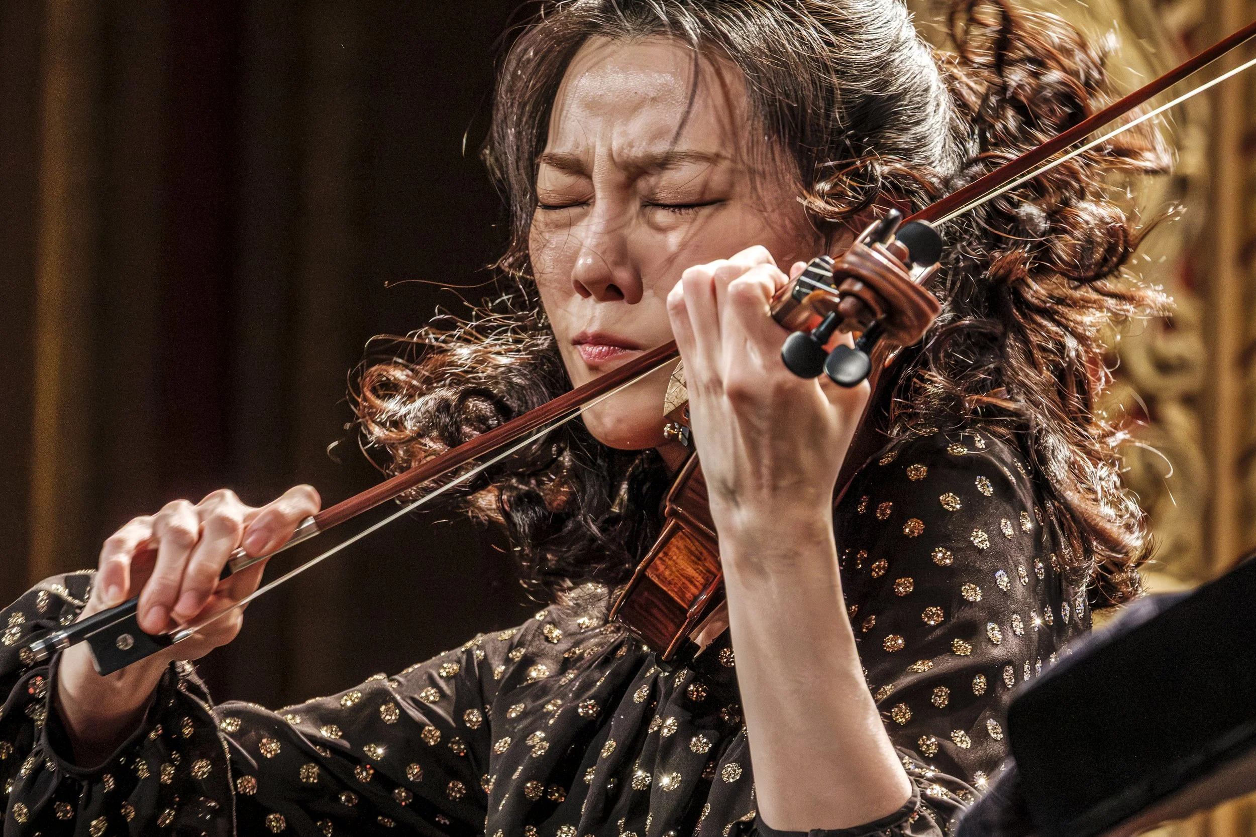 A close up photo of volinist Clara-Jumi Kang playing her violin with her hair whipping around her face.
