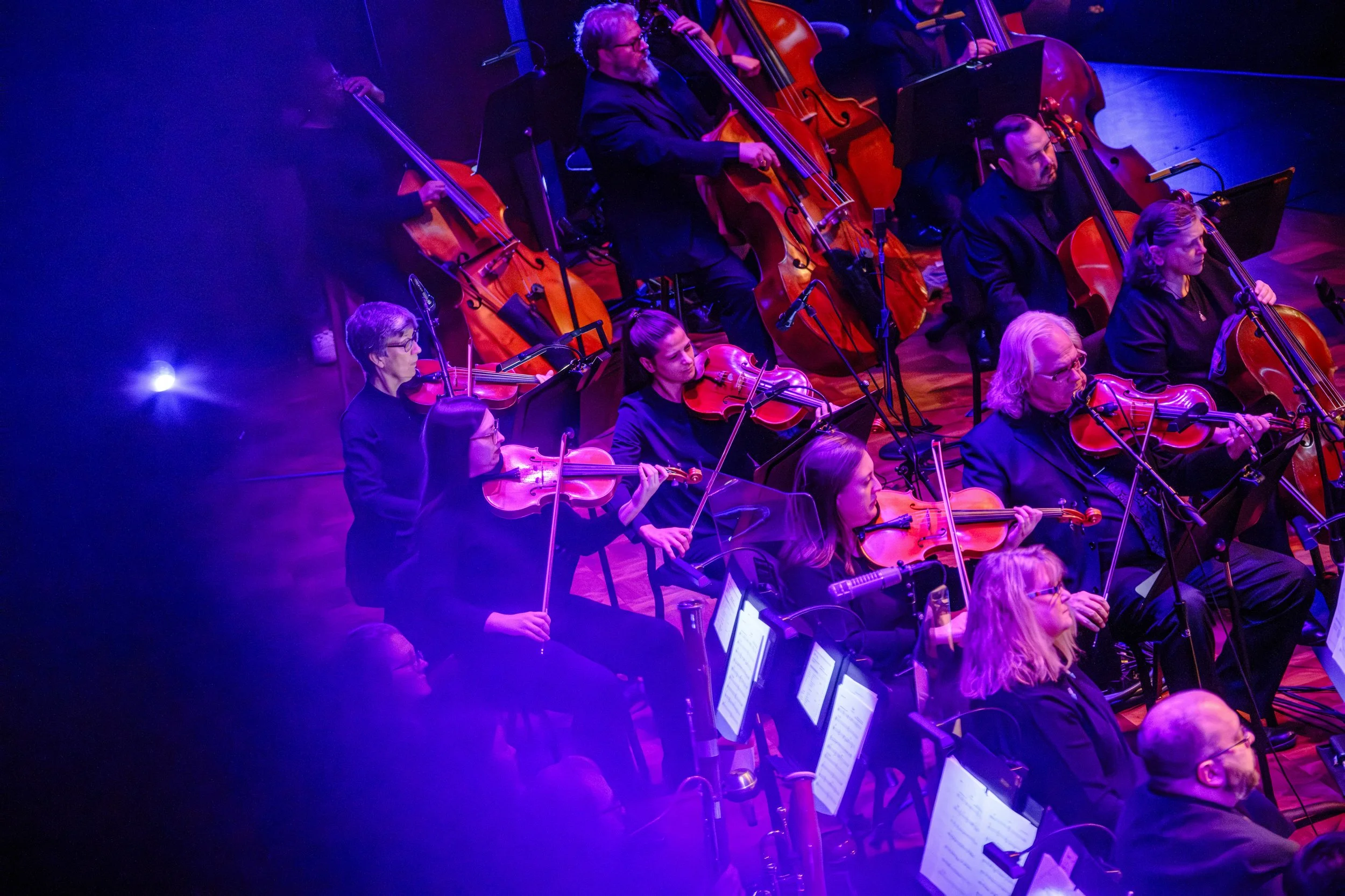 A group of violinists and cellists play with their instruments while bathed in purple stage lighting.