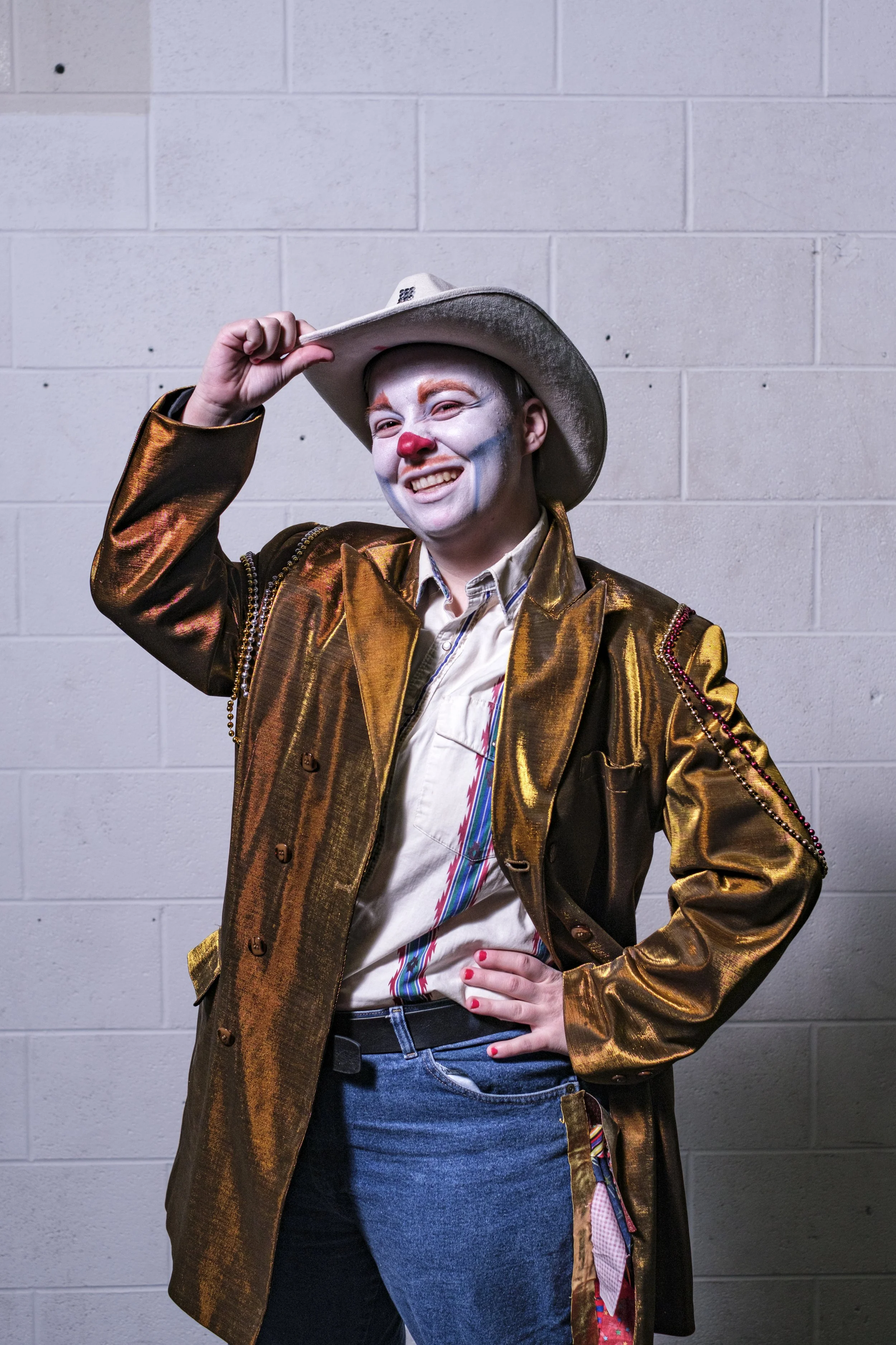 A person wearing clown makeup smiles while tipping their cowboy hat in front of a concrete wall.