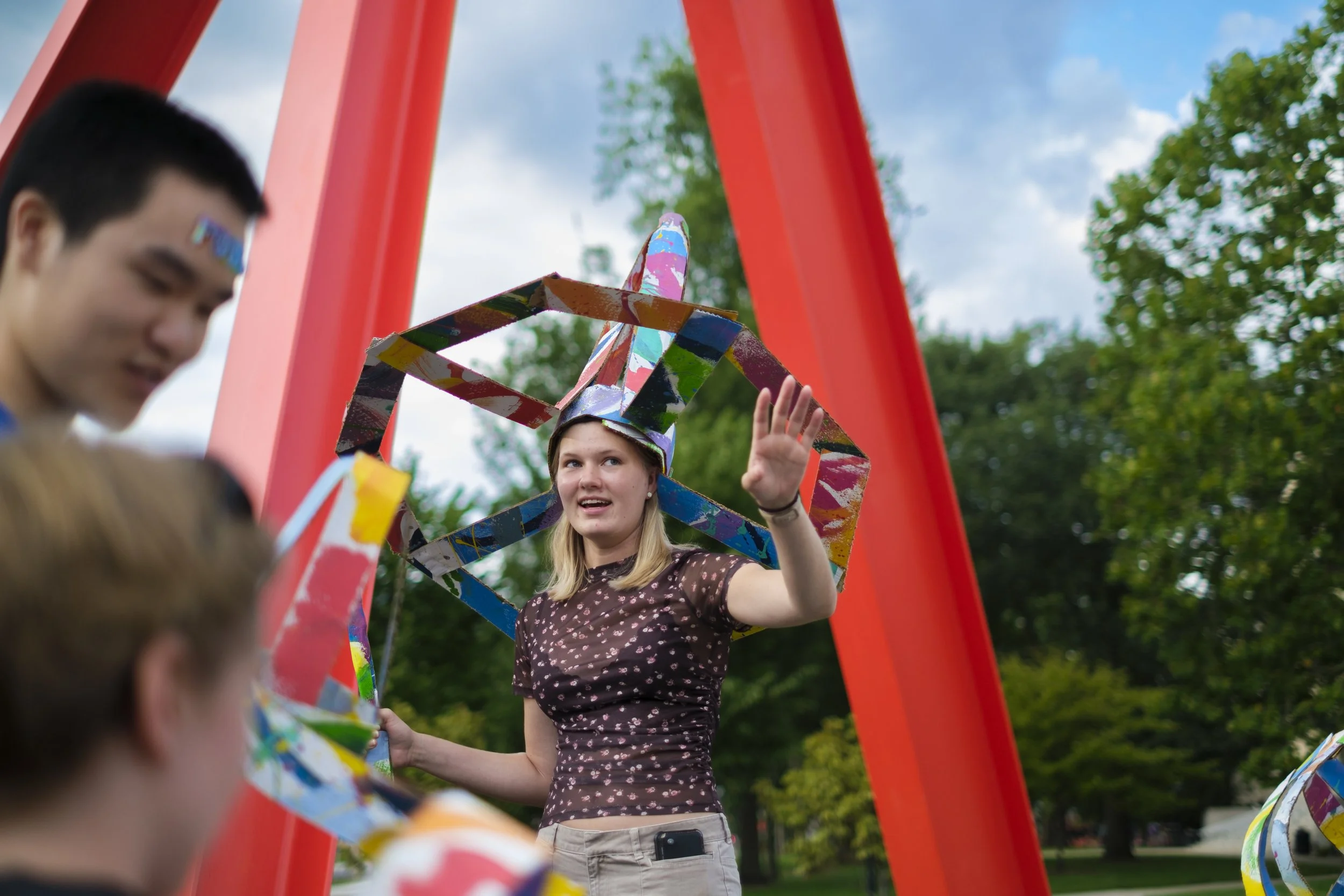 A young woman wearing a jester hat made out of colorful cardboard talks to students during artscapade at the University of Michigan.