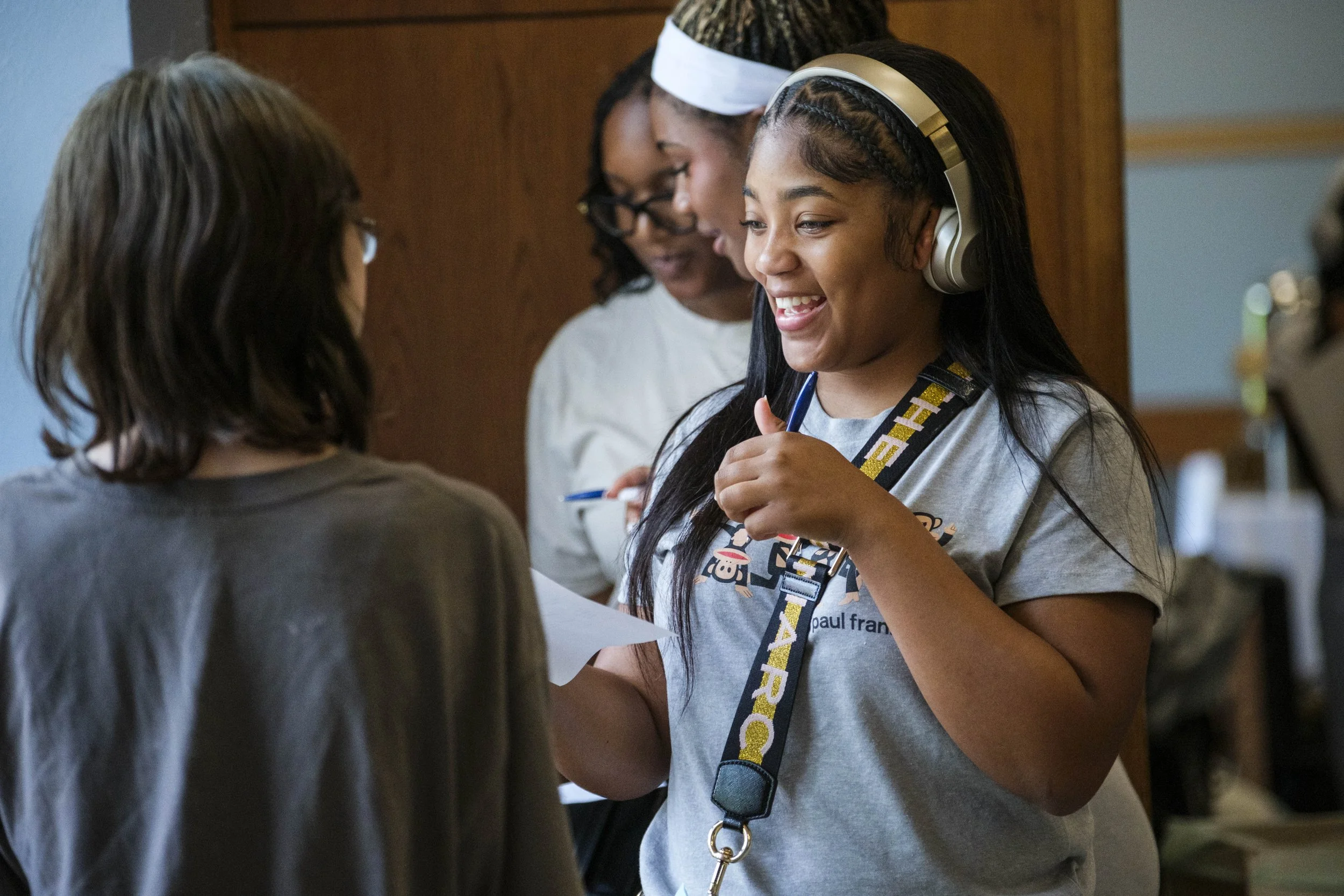 A high school student wearing headphones talks to another student while smiling.
