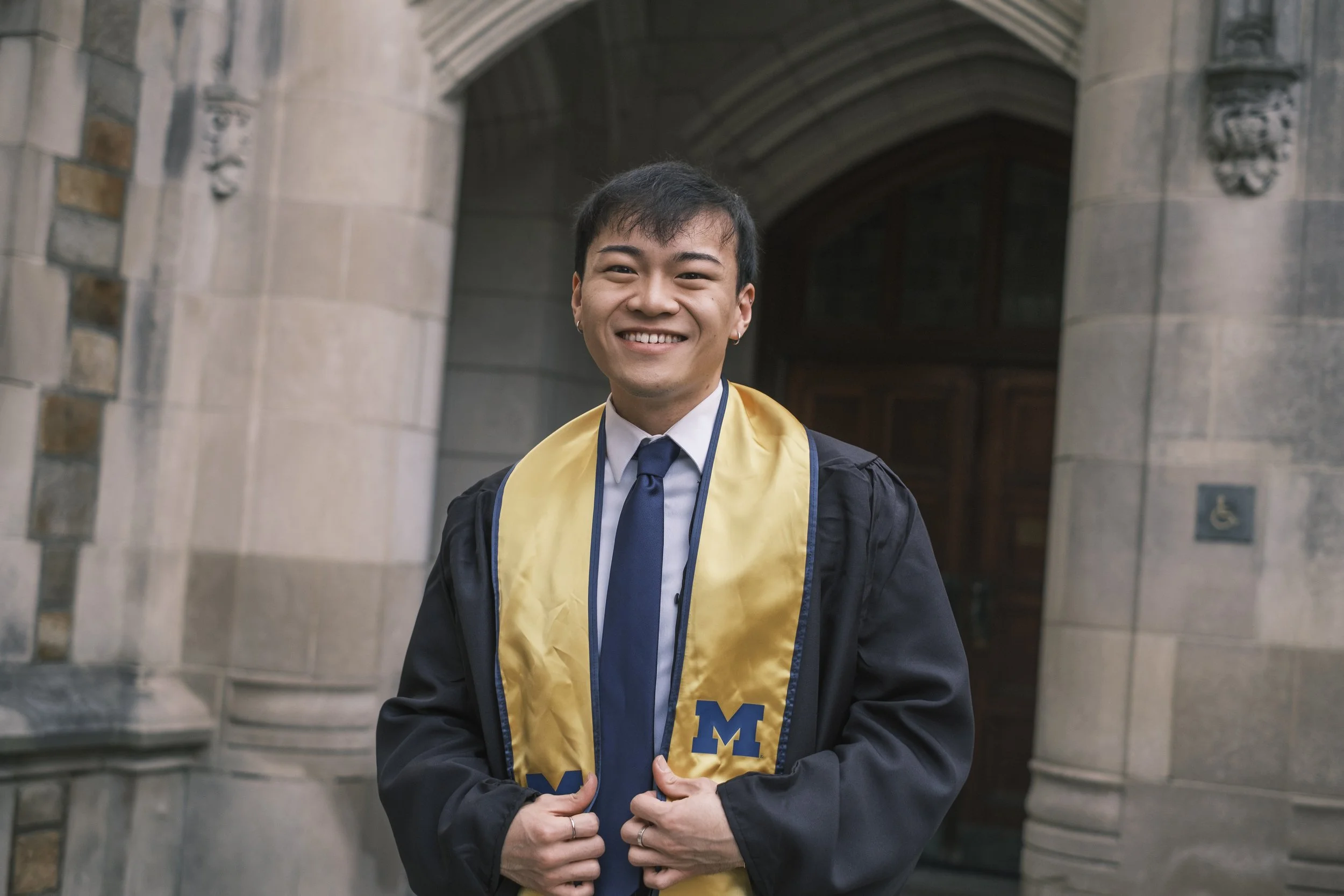 A person smile while holding a golden colored sash while wearing a dress shirt and tie in front of a brick building.