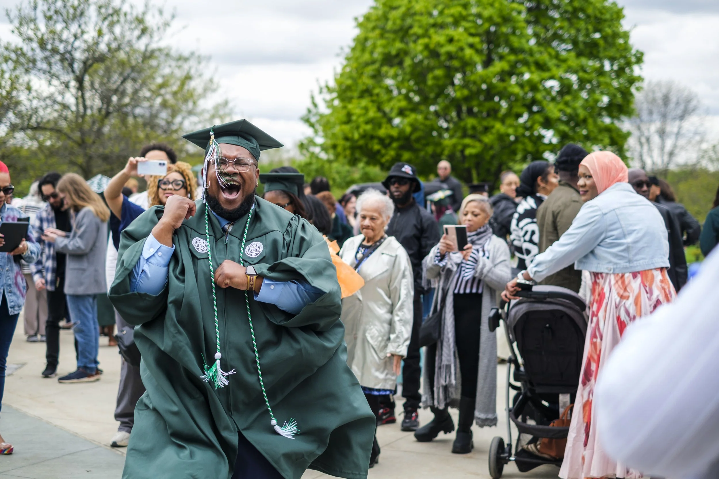 A graduate wearing their graduation cap, gown, and tassels, dances and sings outdoors, surrounded by a crowd of people