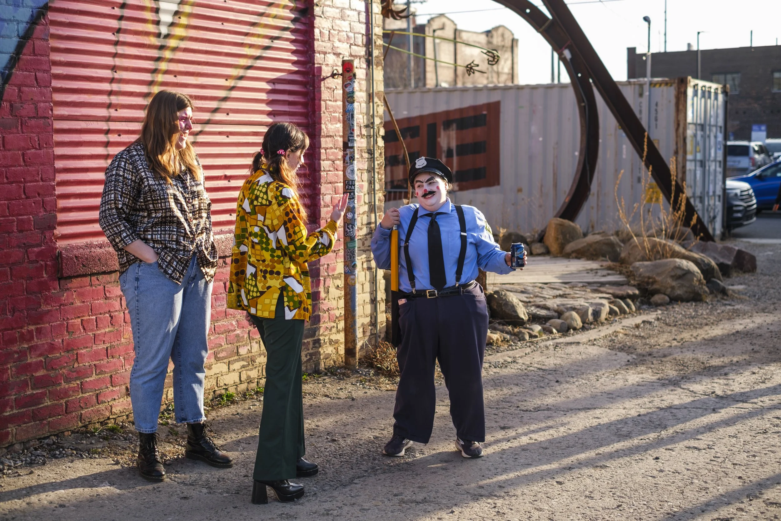 Three people stand outside of a building while the sunsets. One individual is clown wrestler Burt Simmons, in their cop uniform.
