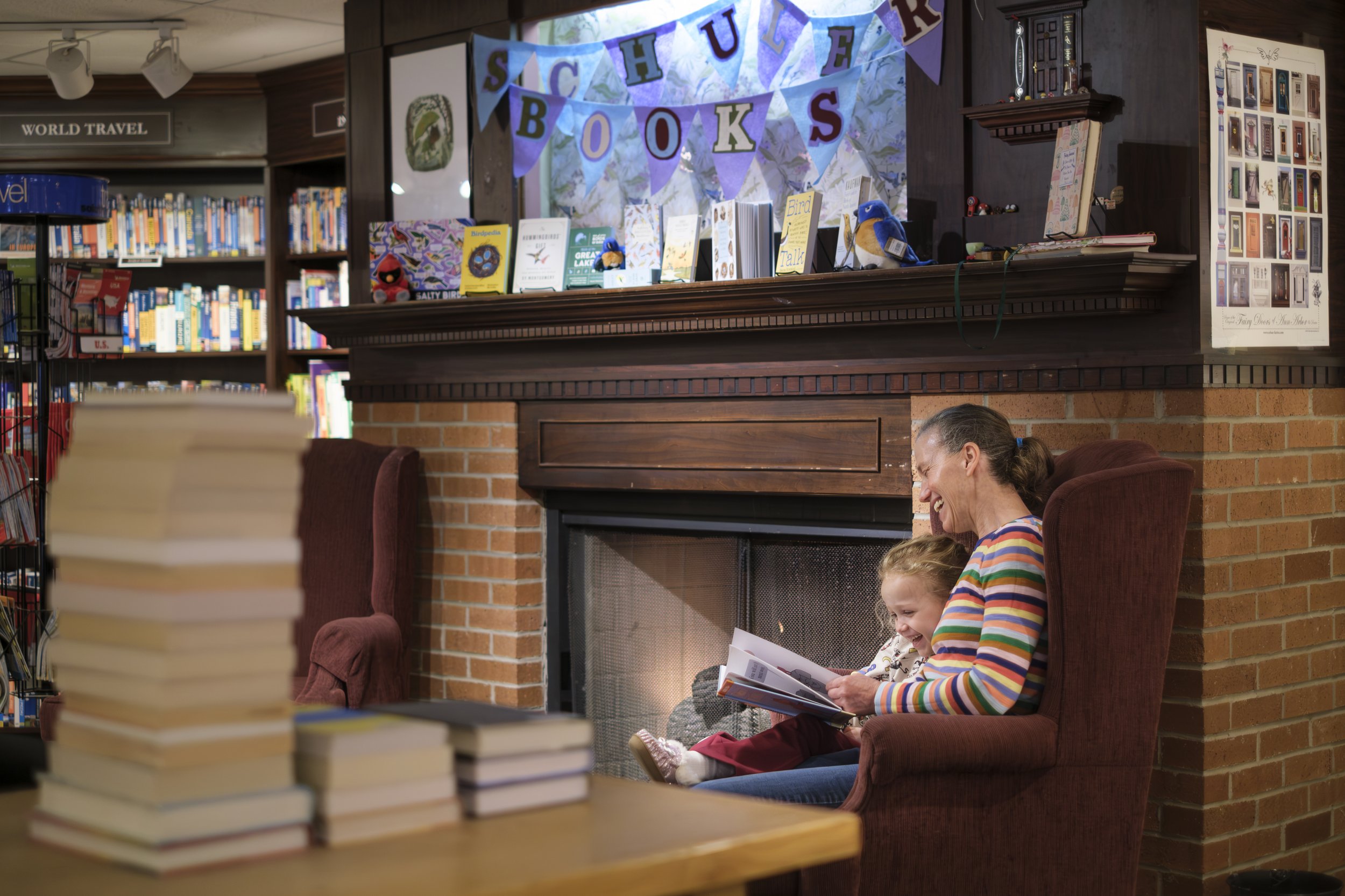 A woman and a young child sit in front of a fireplace reading a book with a banner above them that reads "Schuler Books".