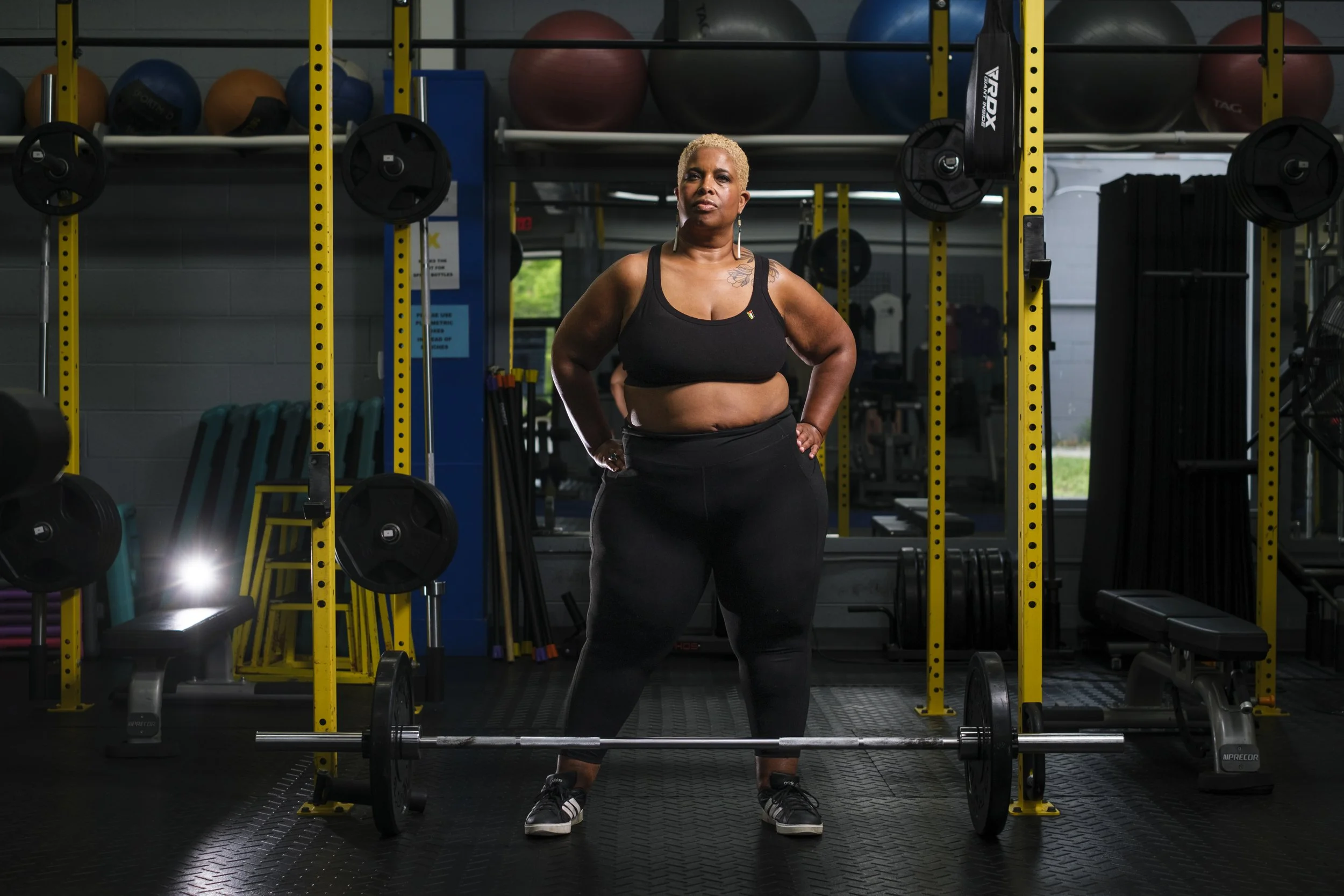 A woman stands confident wither her hands on her hips while wearing a black workout outfit in front of barbells.