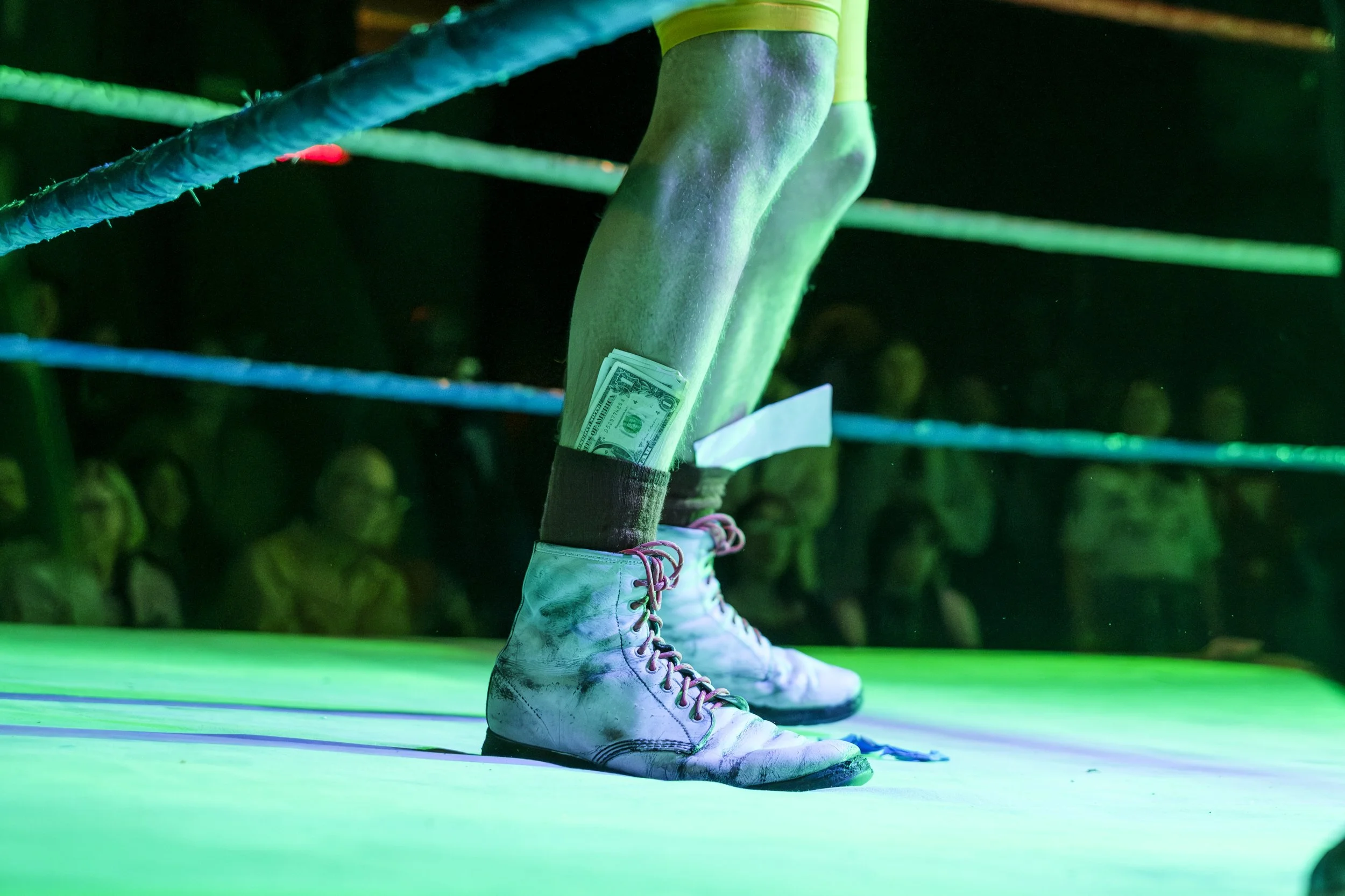 Shoes in a wrestling ring, illuminated by green stage lighting, with dollar bills sticking out of the tops of the shoes.