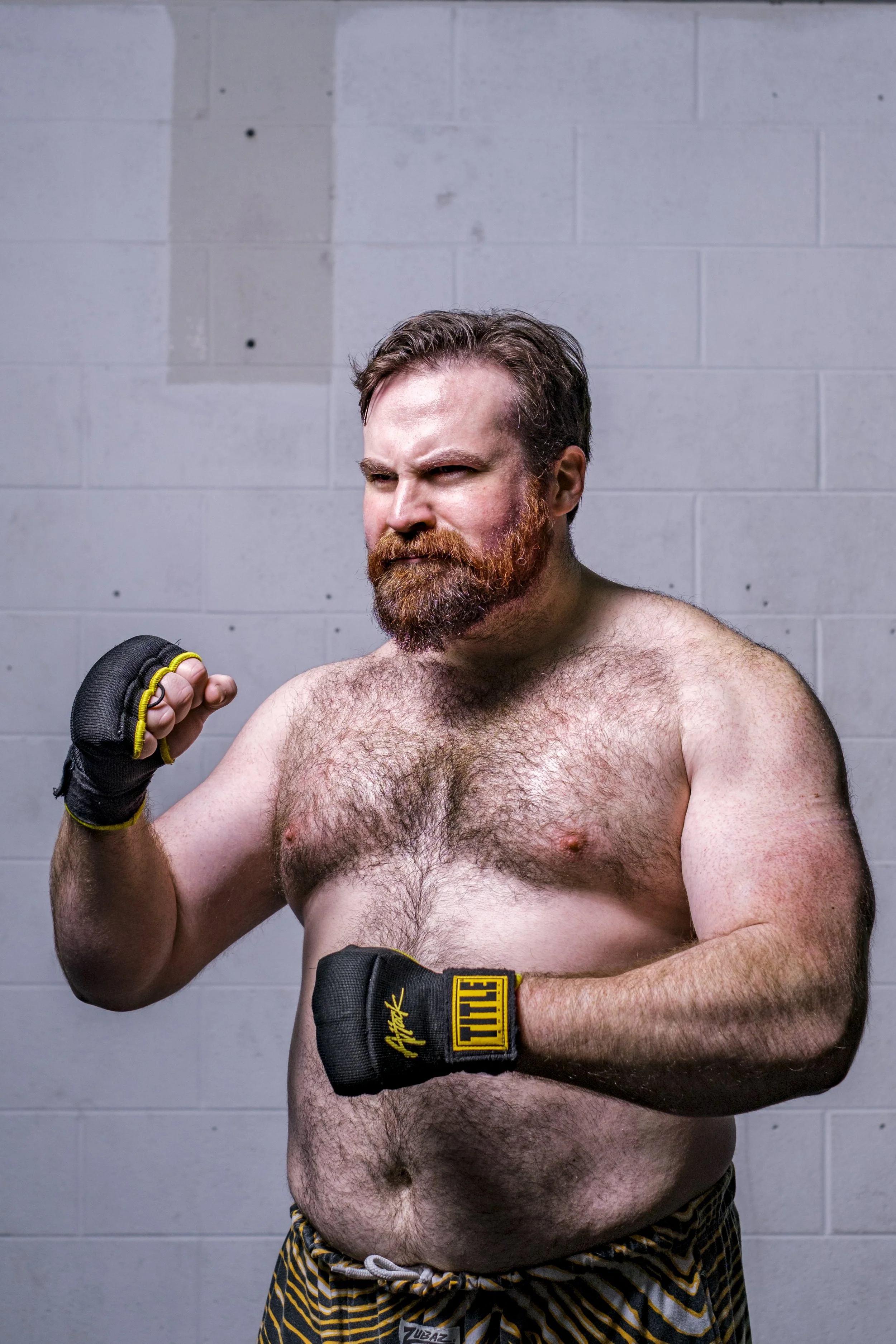 A man with a beard stands in front of a concrete wall wearing TITLE wrestling gloves.