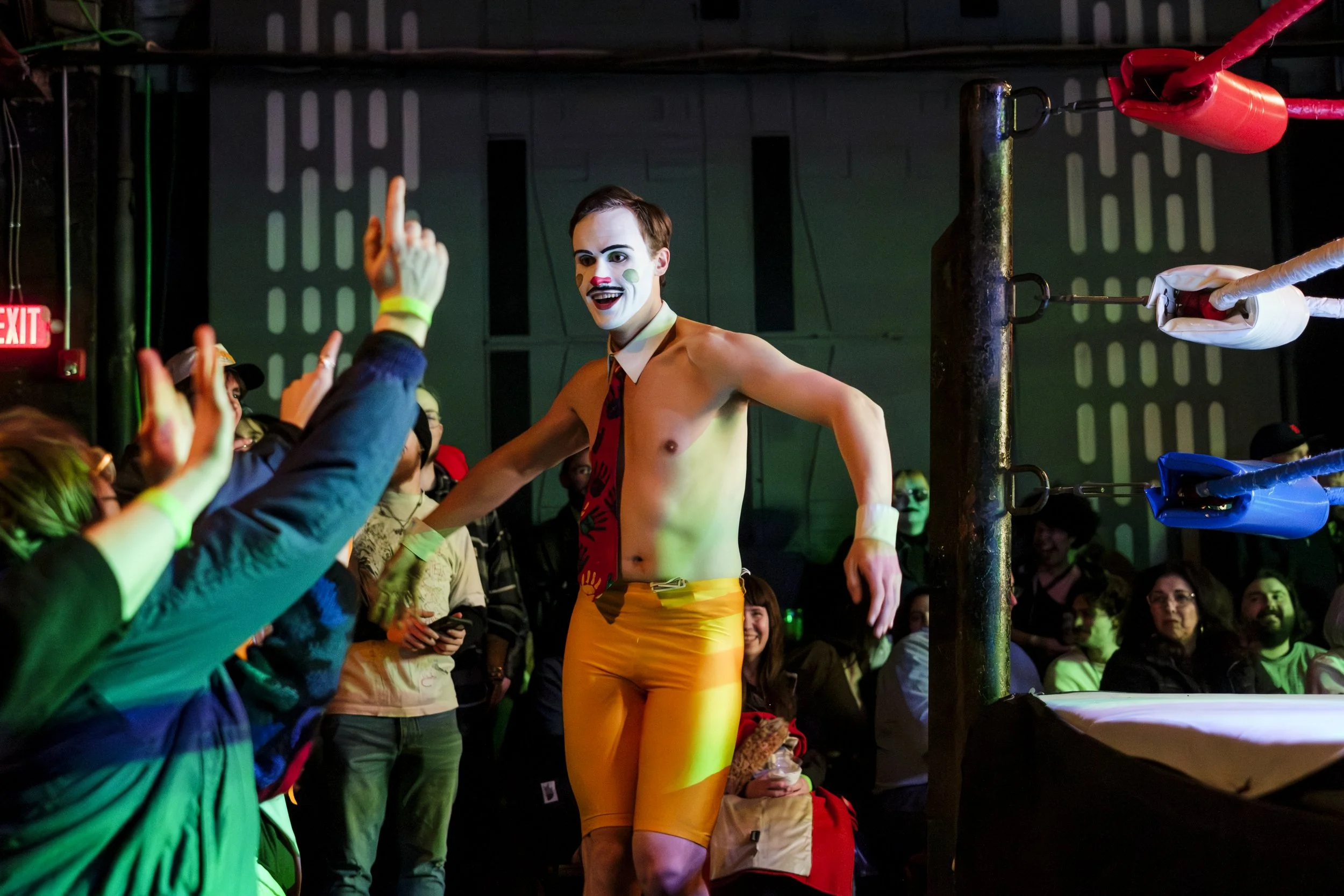 A clown wrestler, known as Generous James, runs around the outside of a wrestling ring, while high-fiving audience members.