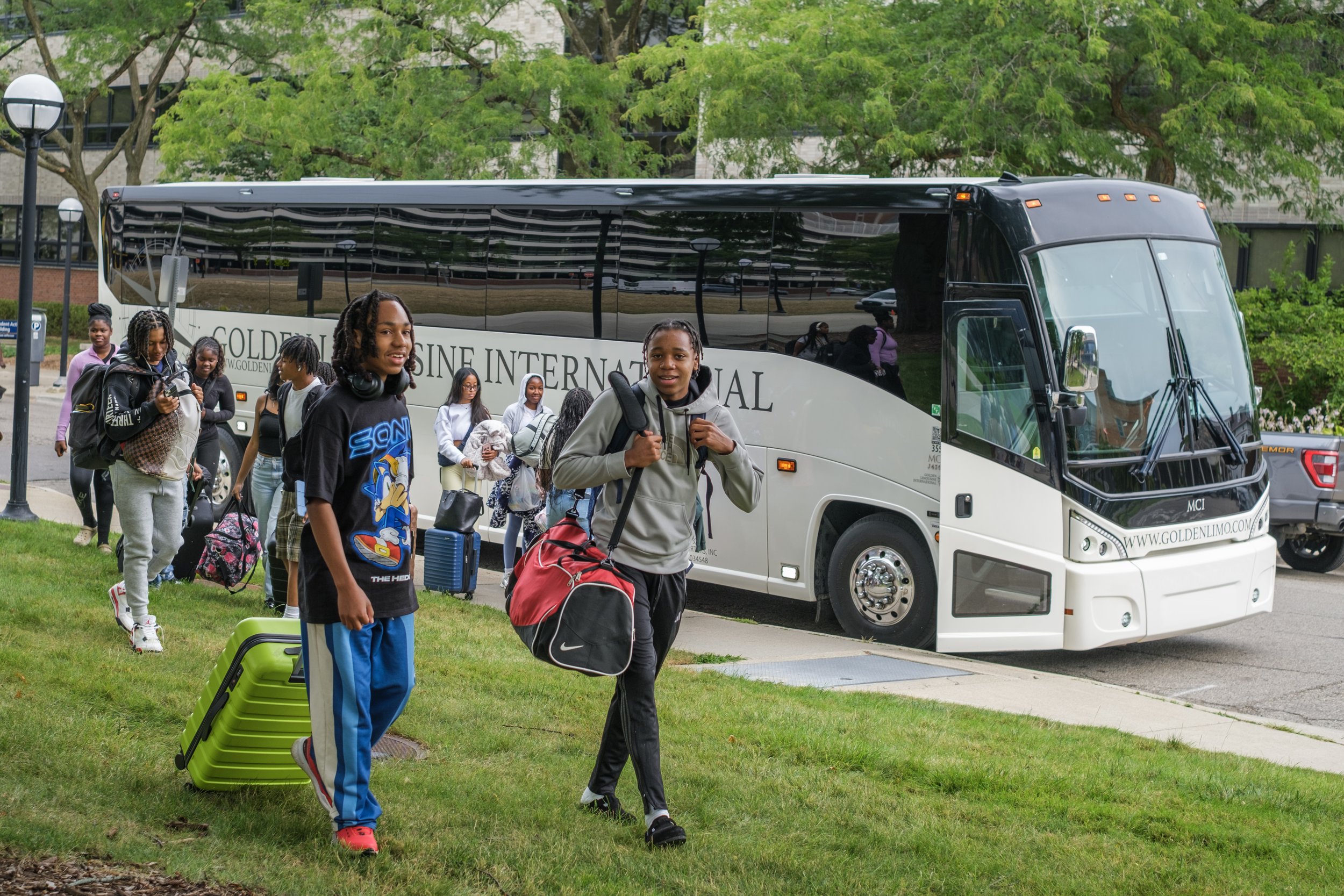Two male high school students walk on grass with a suit case and duffle bags while a large bus with more students grabbing overnight bags in the background.