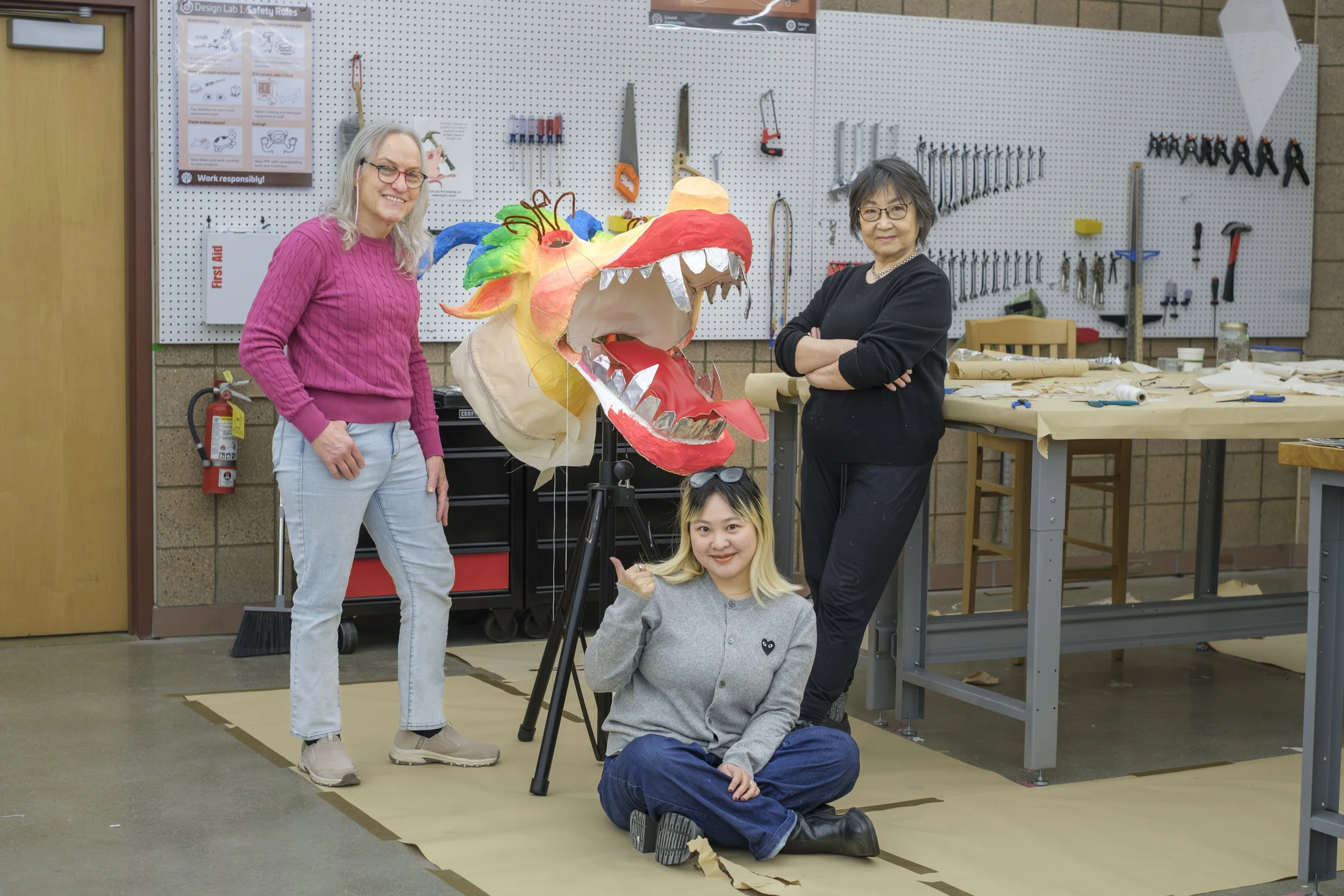 Three people, including artist Zhen Guo, stand and smile next to a color dragon head made out of rice paper in a studio space.