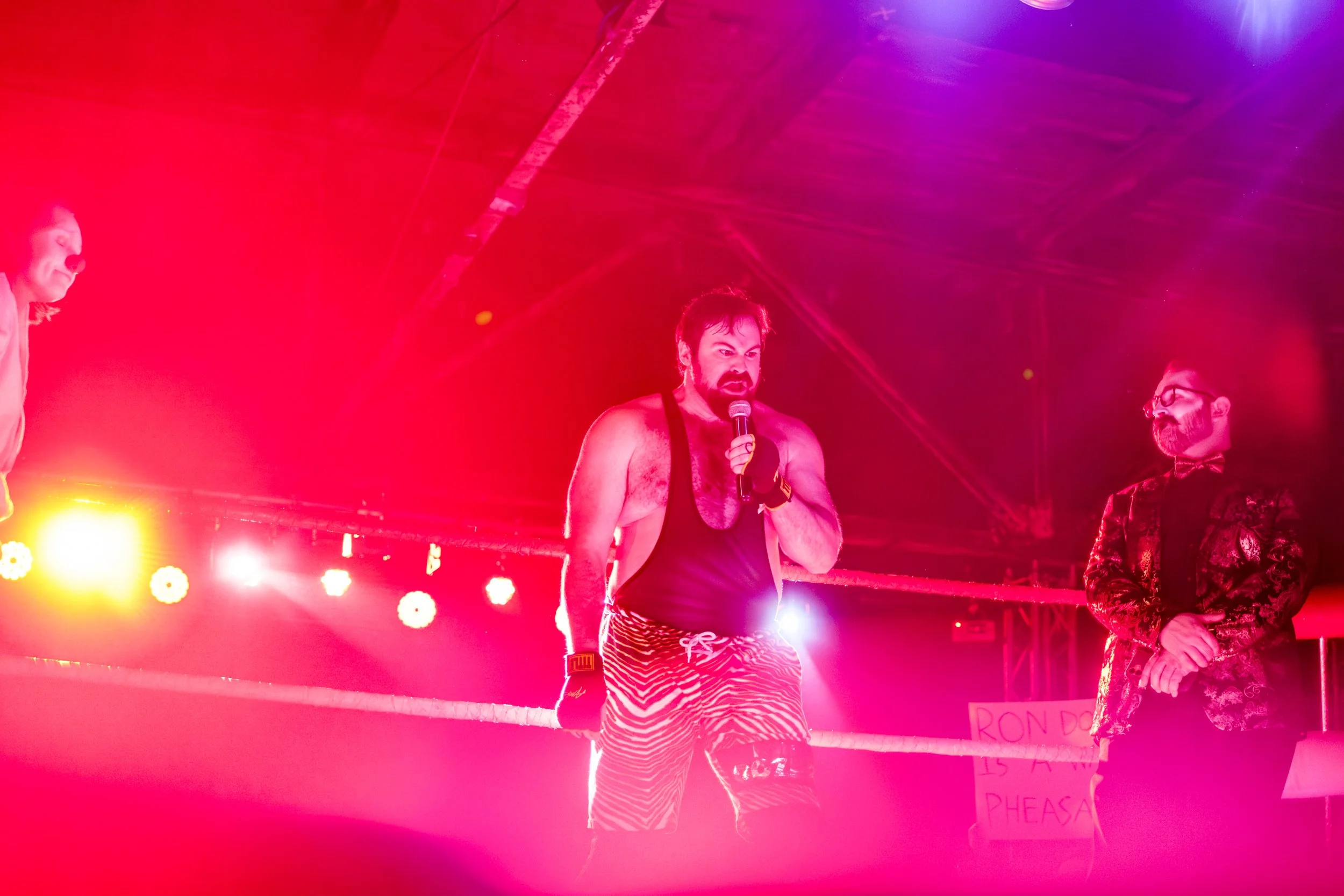 Wrestler Ron Dozer yells into a microphone while in a wrestling ring with referee standing next to him. Red and orange lighting cover the stage.