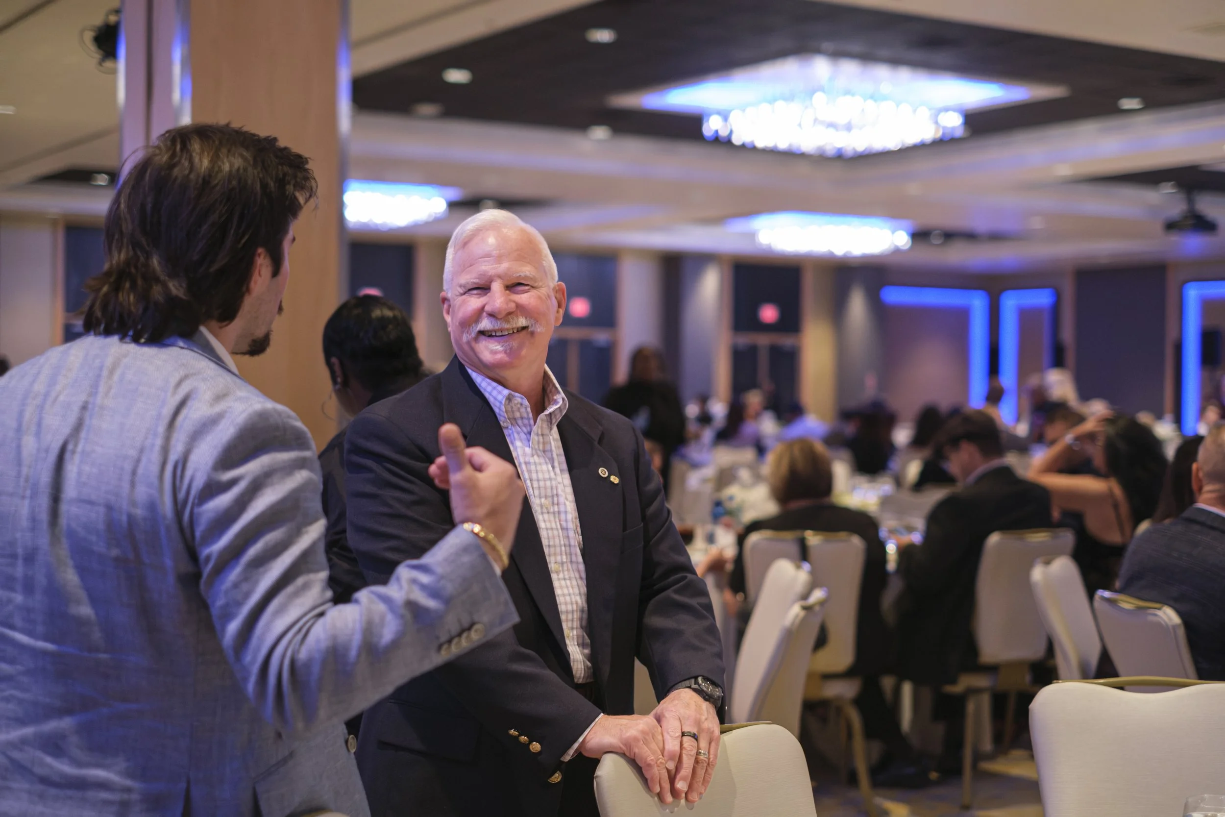 A man wearing a suit with white facial hair smiles while talking to another man wearing a suit in a dining hall.