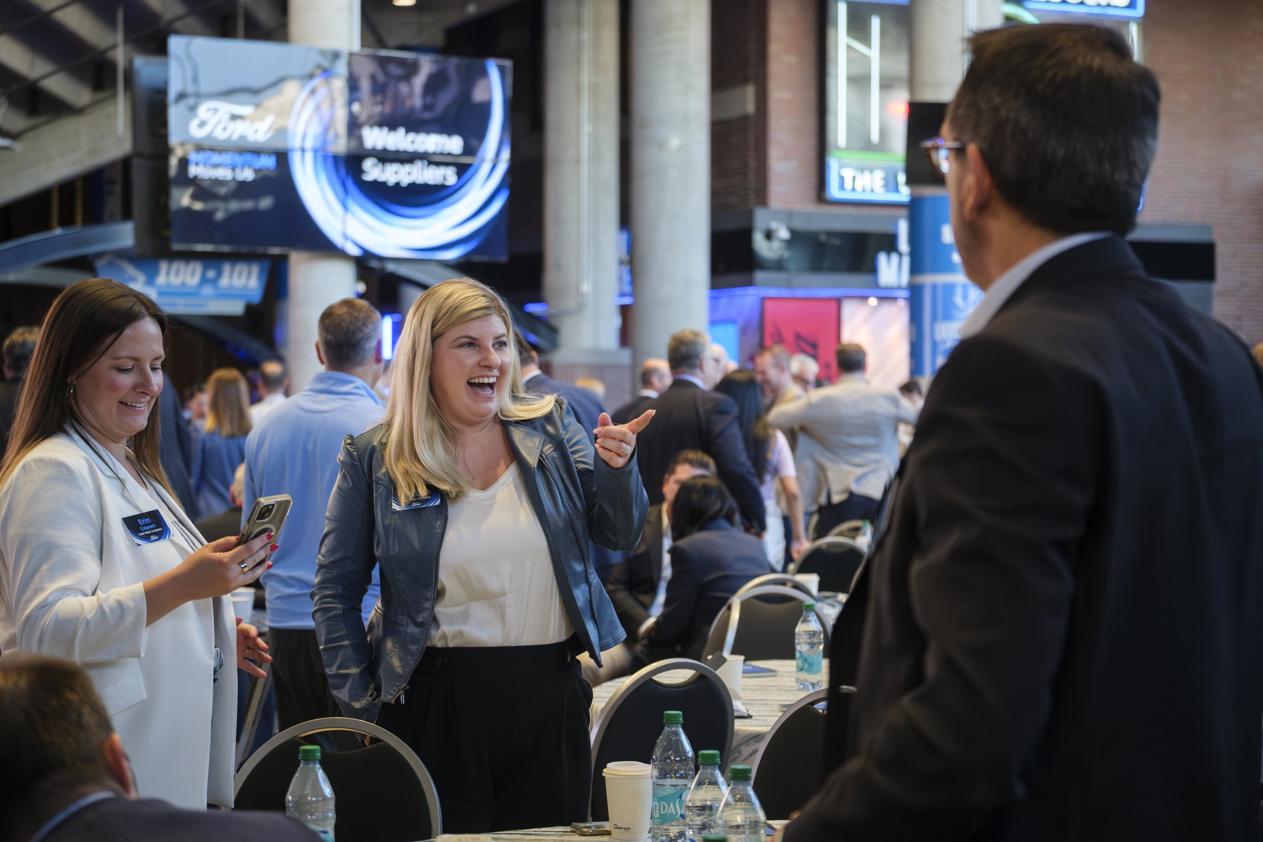 A woman wearing a suit smiles and points while another woman stands next to her, smiling, while looking at her phone.