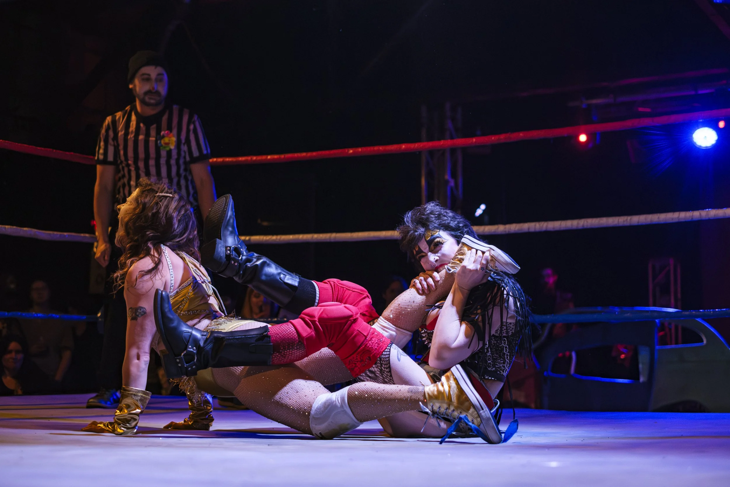 One clown wrestler, known as Pam Hatchett, gnaws at the ankle of Bella Benevolence, another clown wrestler, inside of a wrestling ring.