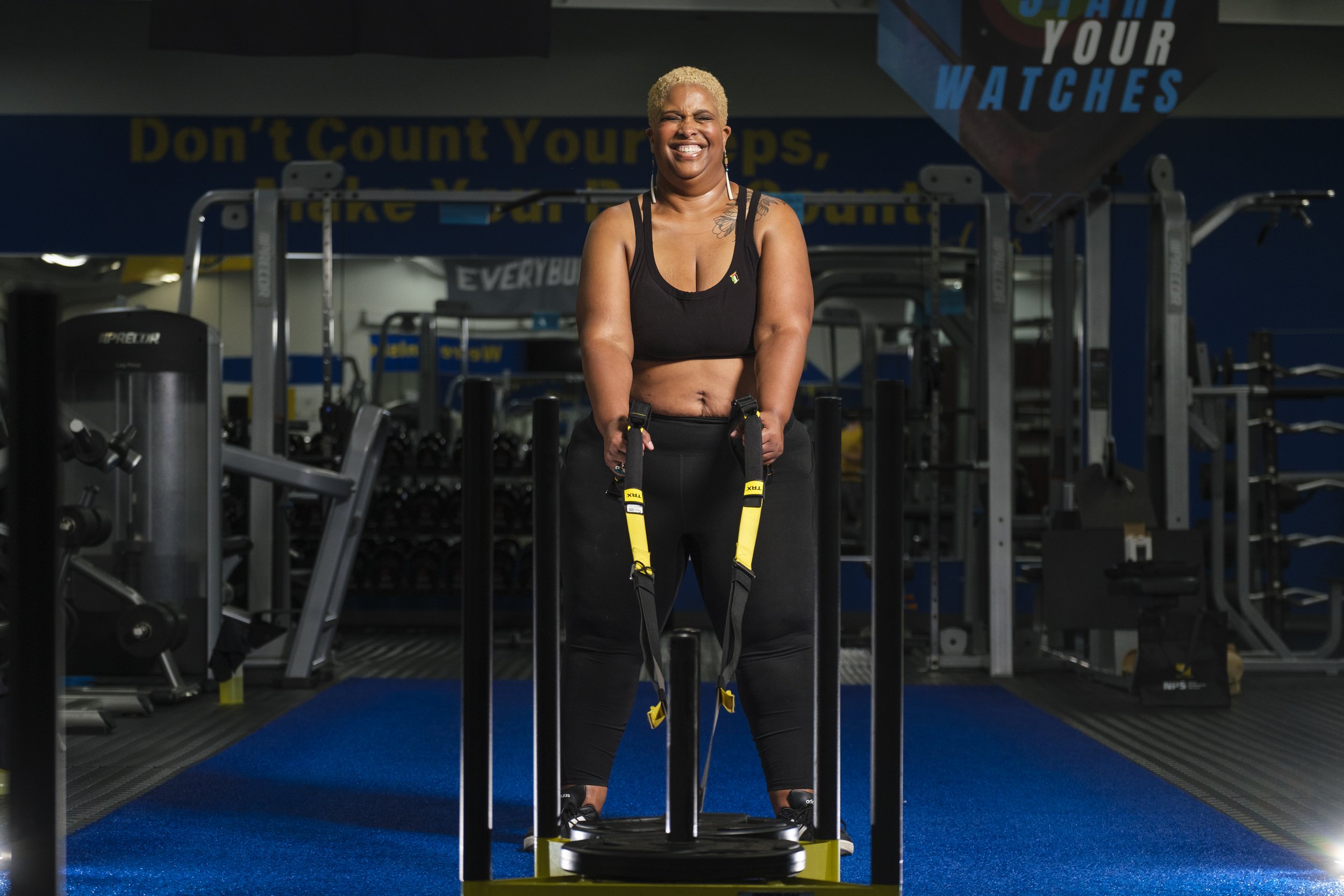 A woman smiles while using a weightlifting machine and ropes to pull it toward her during a weightlifting workout session in a gym.