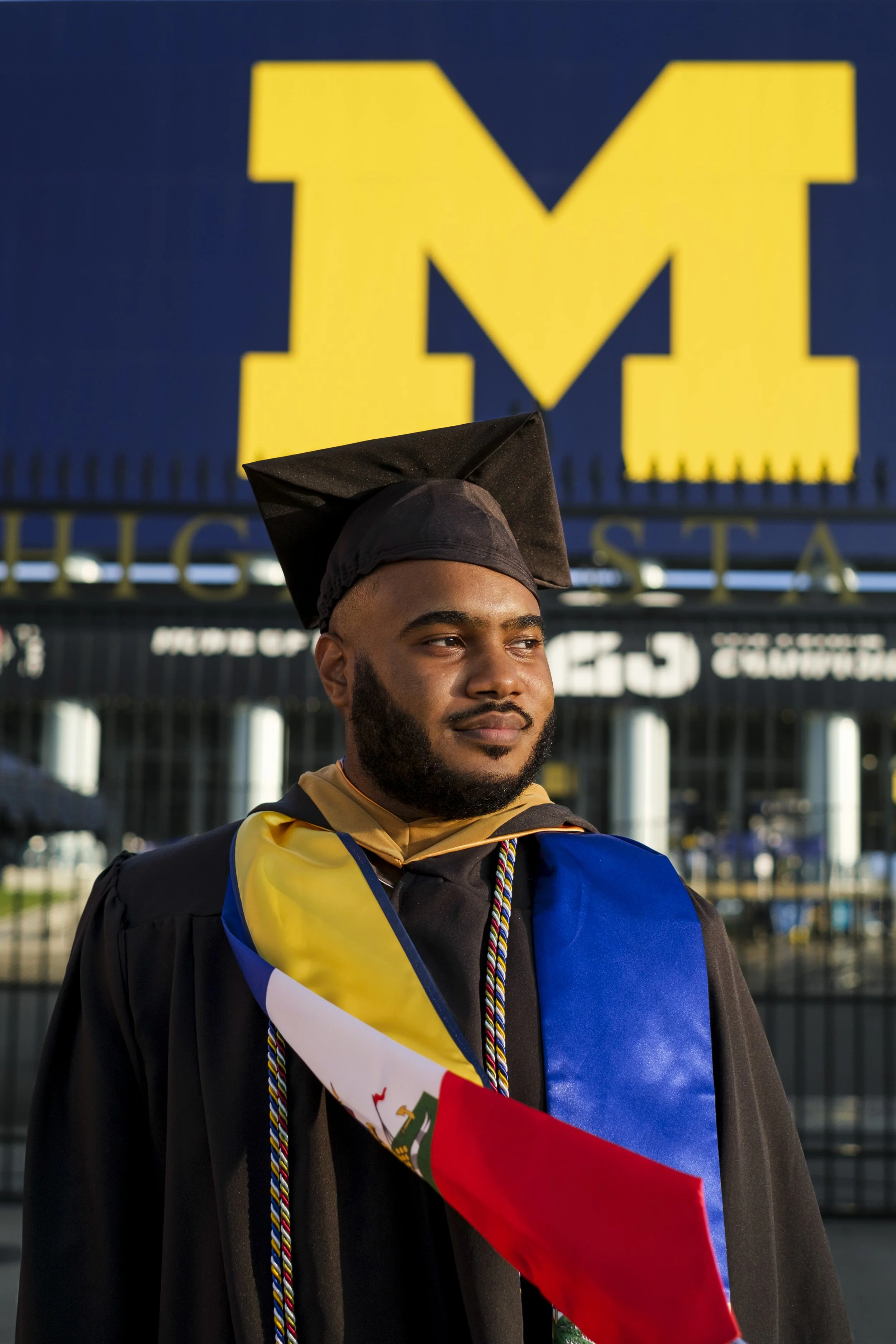 A man wearing his graduation cap, gown, and stoles smiles while looking into the distance with the Block M of Michigan stadium is in the background.