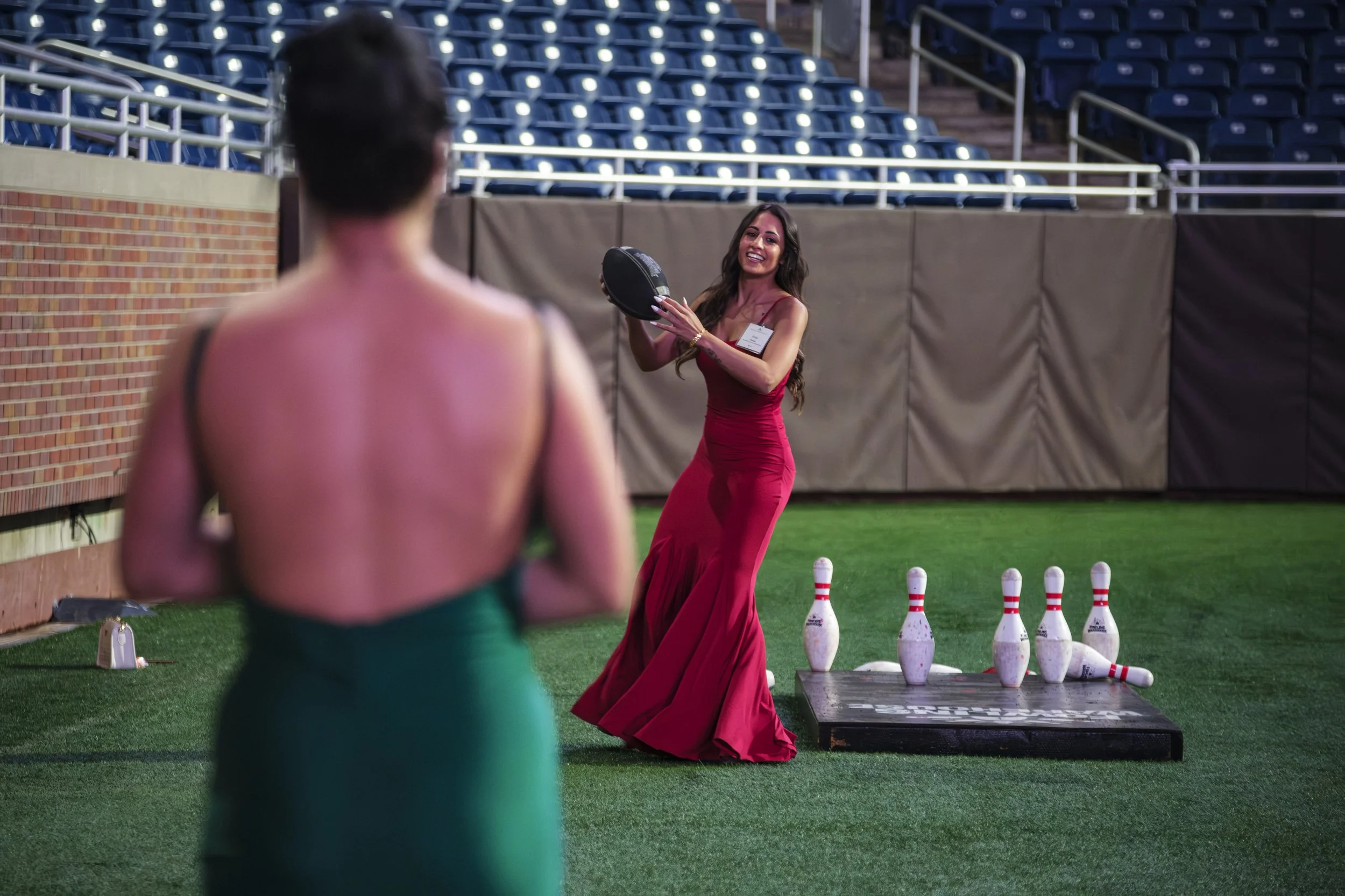 A woman wearing a red dress smiles while holding a football while bowling pins lay on the ground next to her at Ford Field during the Building Bridges Gala.
