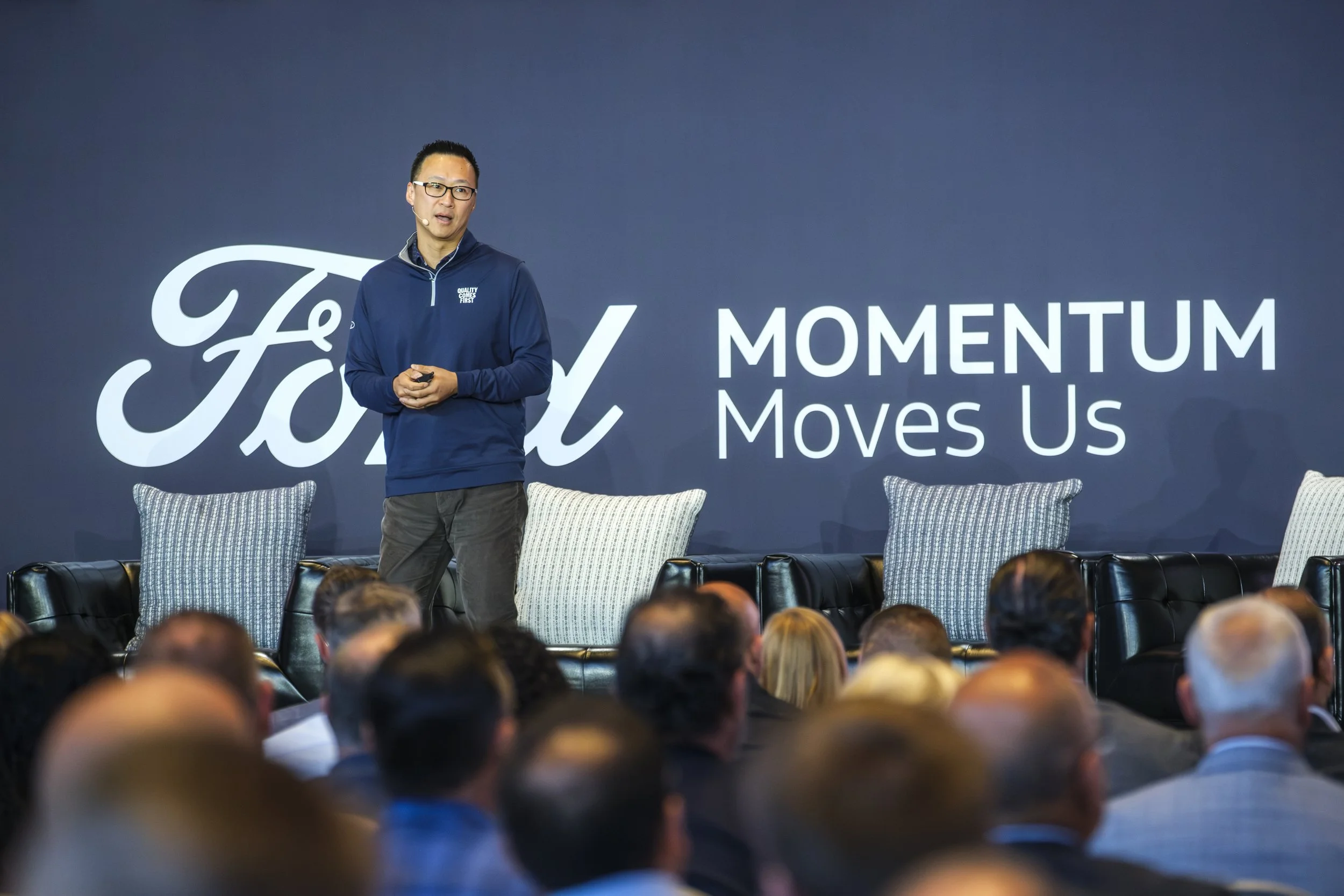 A man wearing glasses stands on stage while holding a clicker and talks to the audience in front of him. In the background is a banner that reads "Ford Momentum Moves Us".