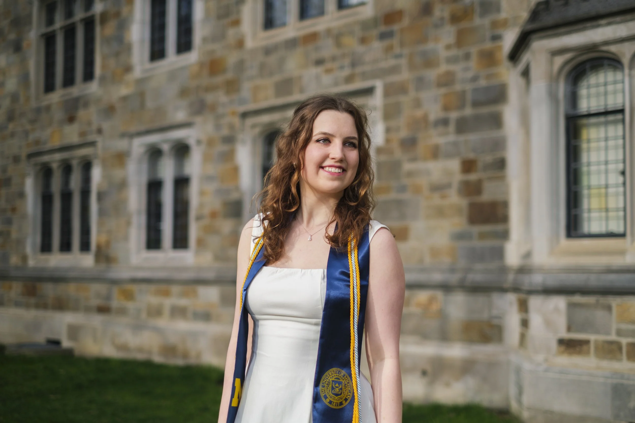 A woman smiles while wearing a dress and a University of Michigan sash and tassels around her shoulders against a brick wall with windows.
