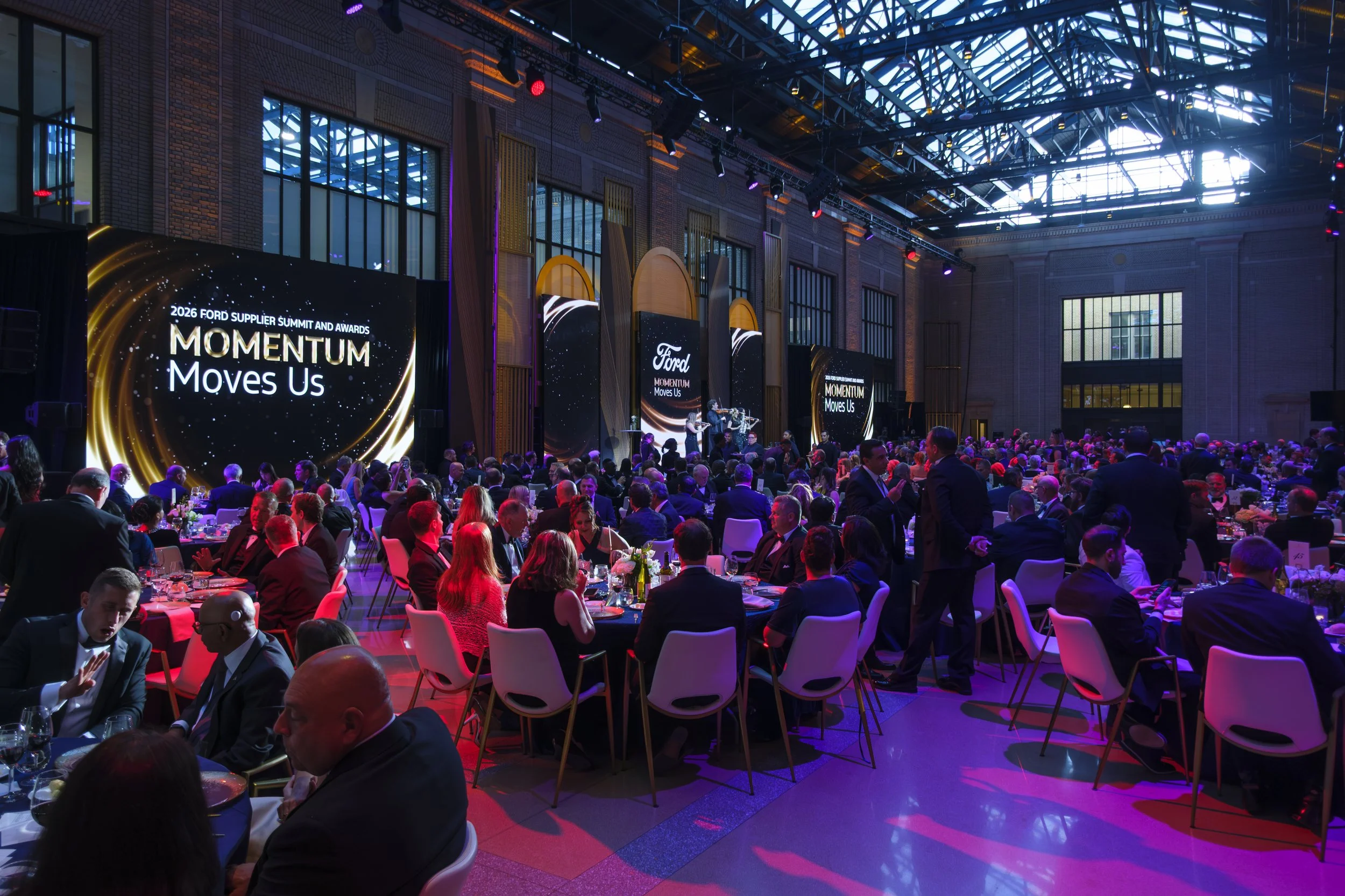 A wide shot showing a room full of people is lit with red and purple lighting with screens in the background that read "2026 Ford Supplier Summit And Awards Momentum Moves Us" at the Ford Supplier Awards at Michigan Central Station in Detroit.