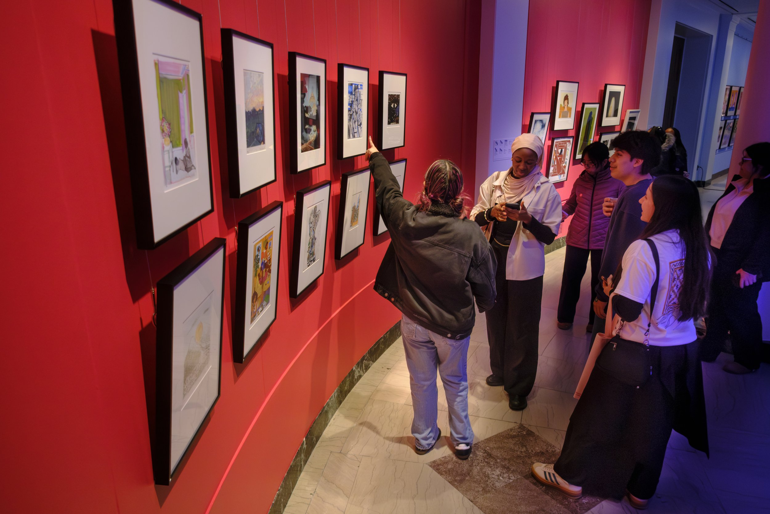 A group of students stand in front of a wall of art while one points at a painting on the wall.