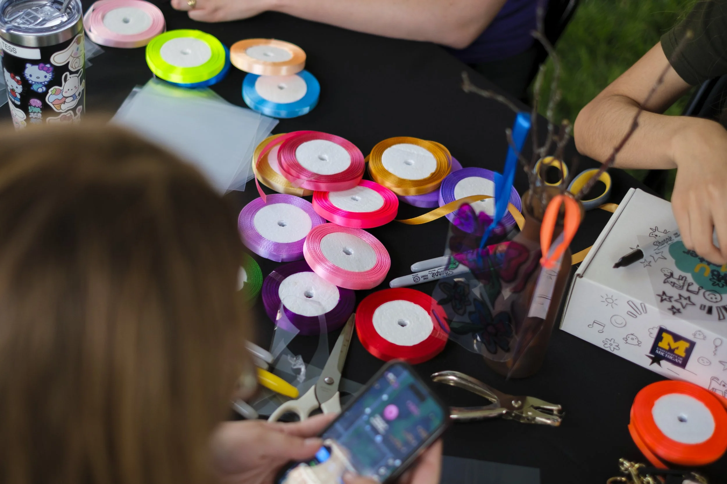 Colorful satin ribbons lay scattered on a table with other art supplies, including a hole puncher and scissors.