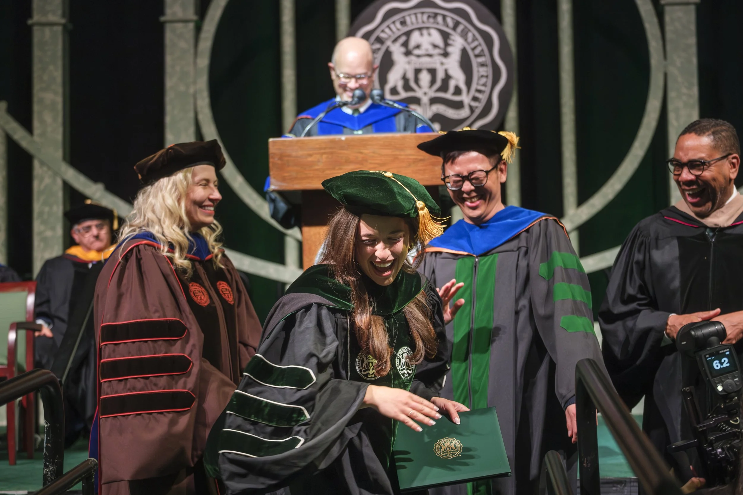 Three people in doctoral regalia smile at one another while a person speaks at a podium and the seal of Eastern Michigan University in the background.