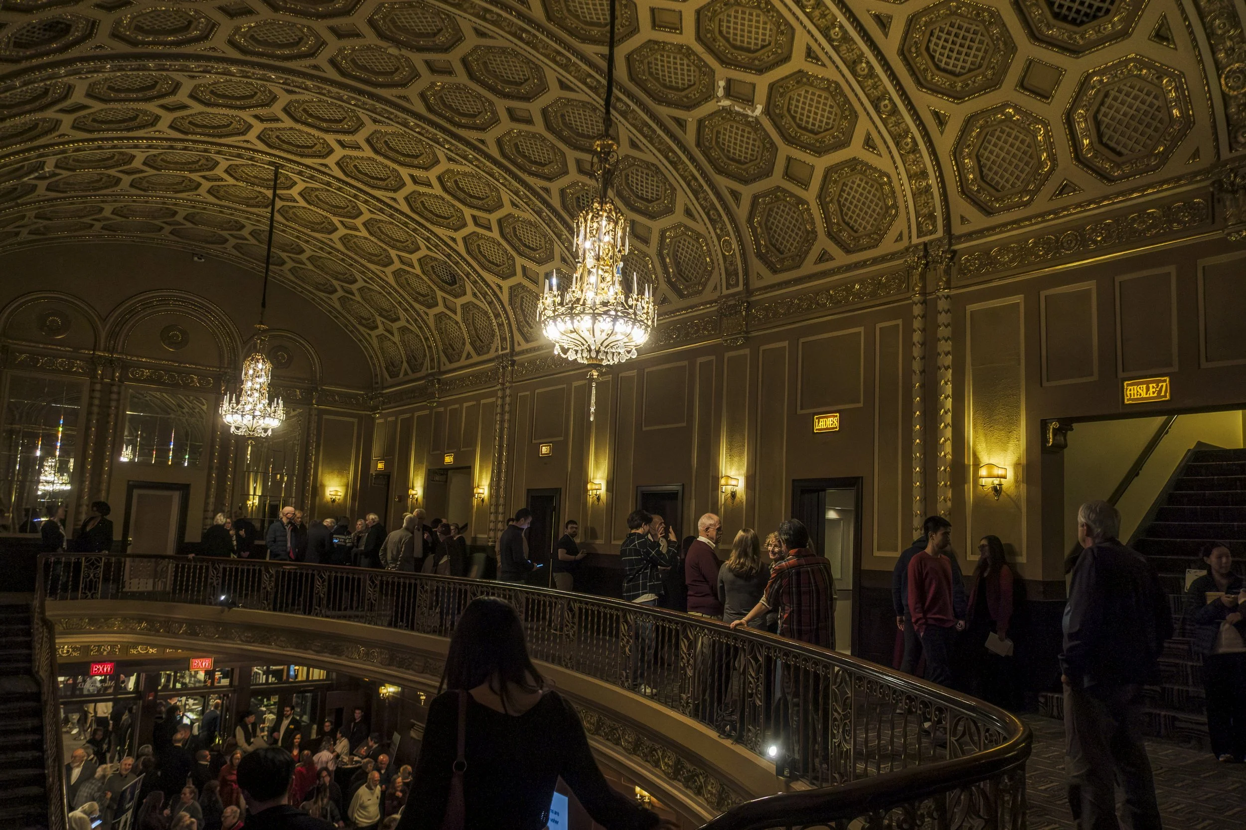 A group of people stand around a balcony in an ornate theater with chandeliers hanging the ceiling.