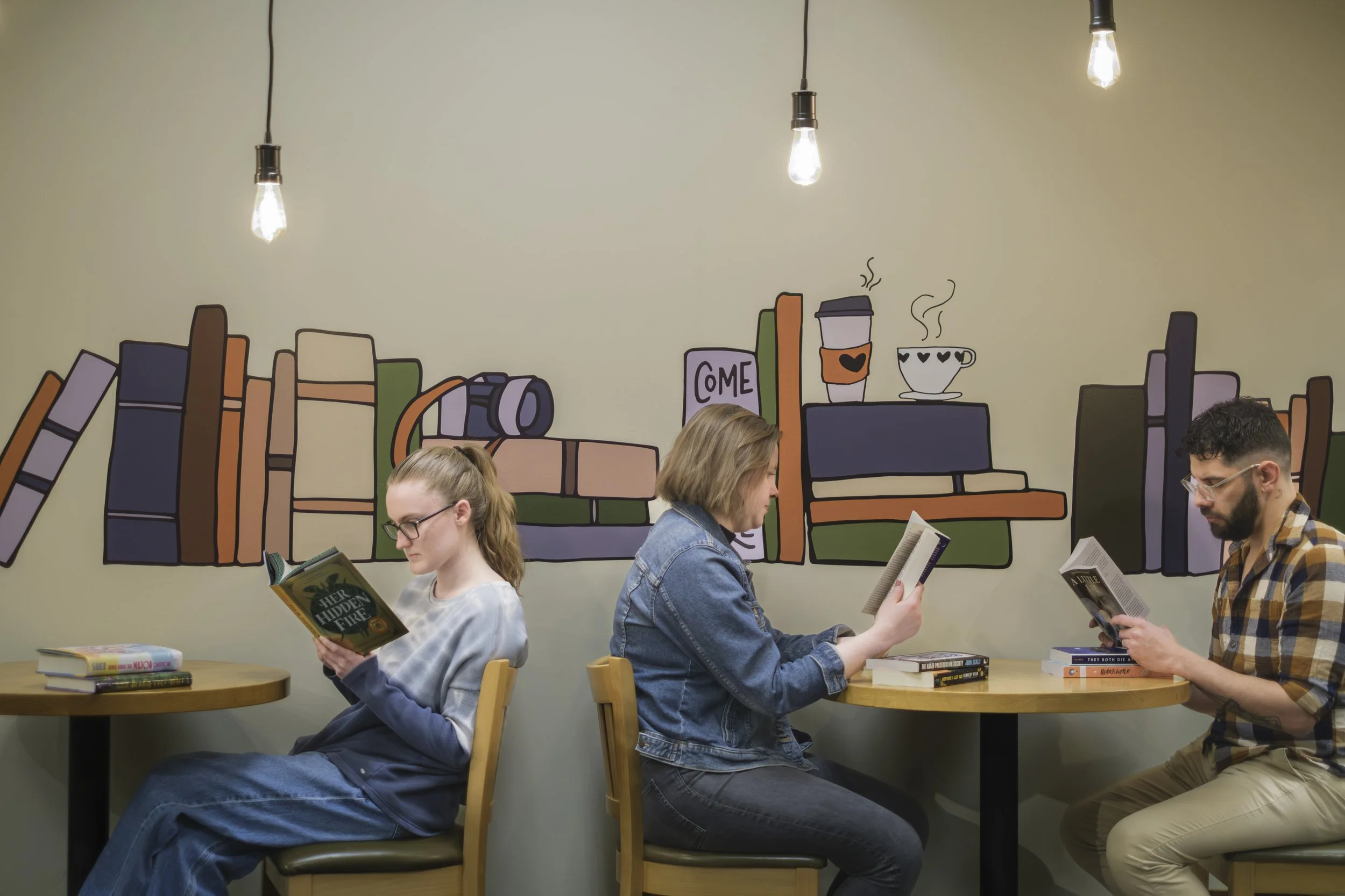 Three people sit at high-top tables reading books with a mural of books and coffee behind them.